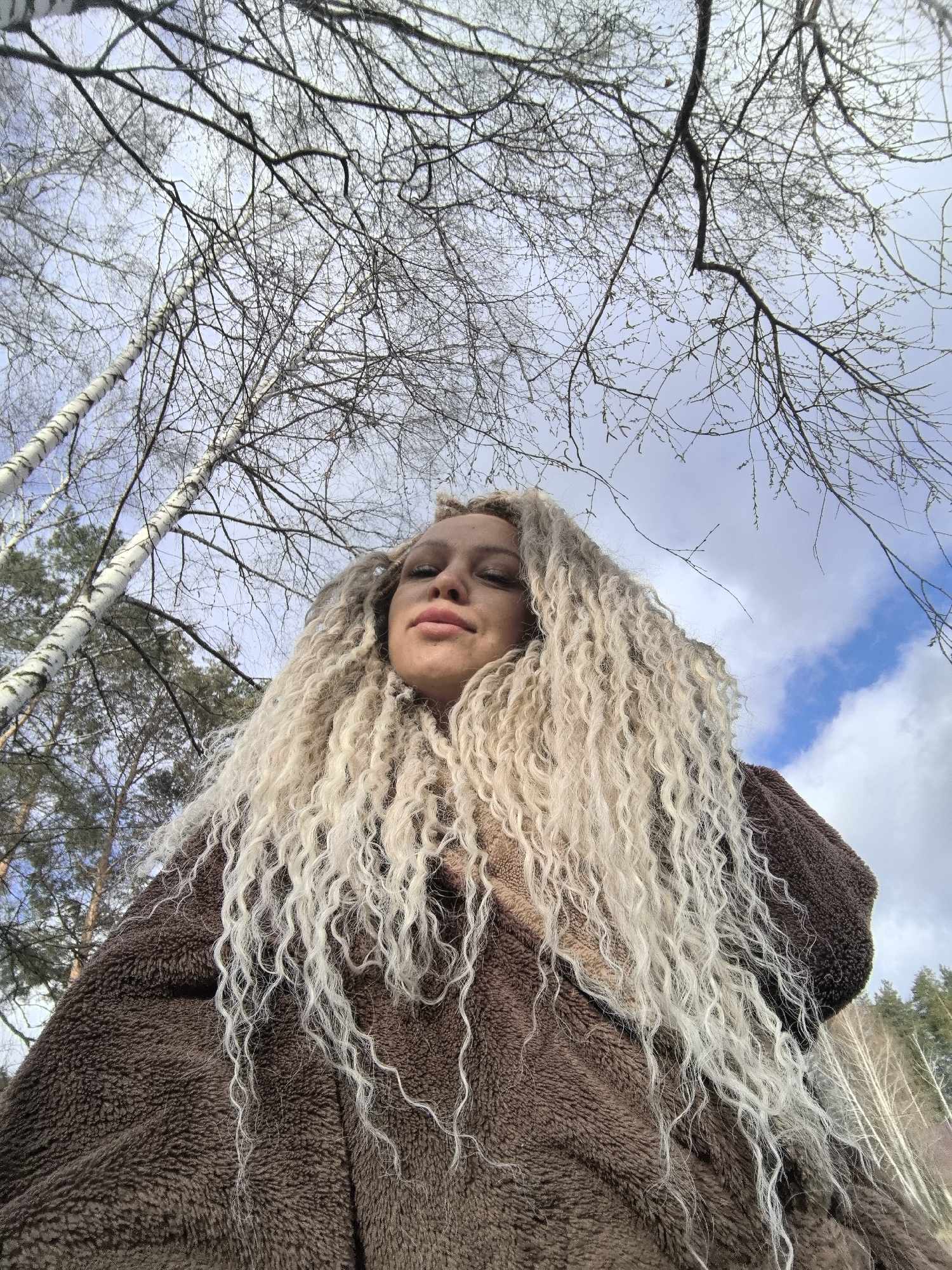 This low-angle outdoor portrait features a woman with voluminous, light blonde or white curly hair, likely dreadlocks or synthetic extensions, cascading over her shoulders and chest. She is wearing a thick, textured, brown, faux-fur or fleece coat. Her face is partially visible, with a calm, slightly pouty expression and full lips.
The setting is a natural, wooded area, characteristic of Noviy, Russia, during late autumn or winter. Bare tree branches, primarily from deciduous trees, are prominent against a light blue sky with scattered white clouds. Several distinct white-barked birch trees with dark markings are visible on the left and center-left of the frame. In the background, darker green coniferous trees can be seen. The woman is looking towards the camera, suggesting she is posing for a selfie or a photograph. The natural light indicates it is daytime. There is no visible text.