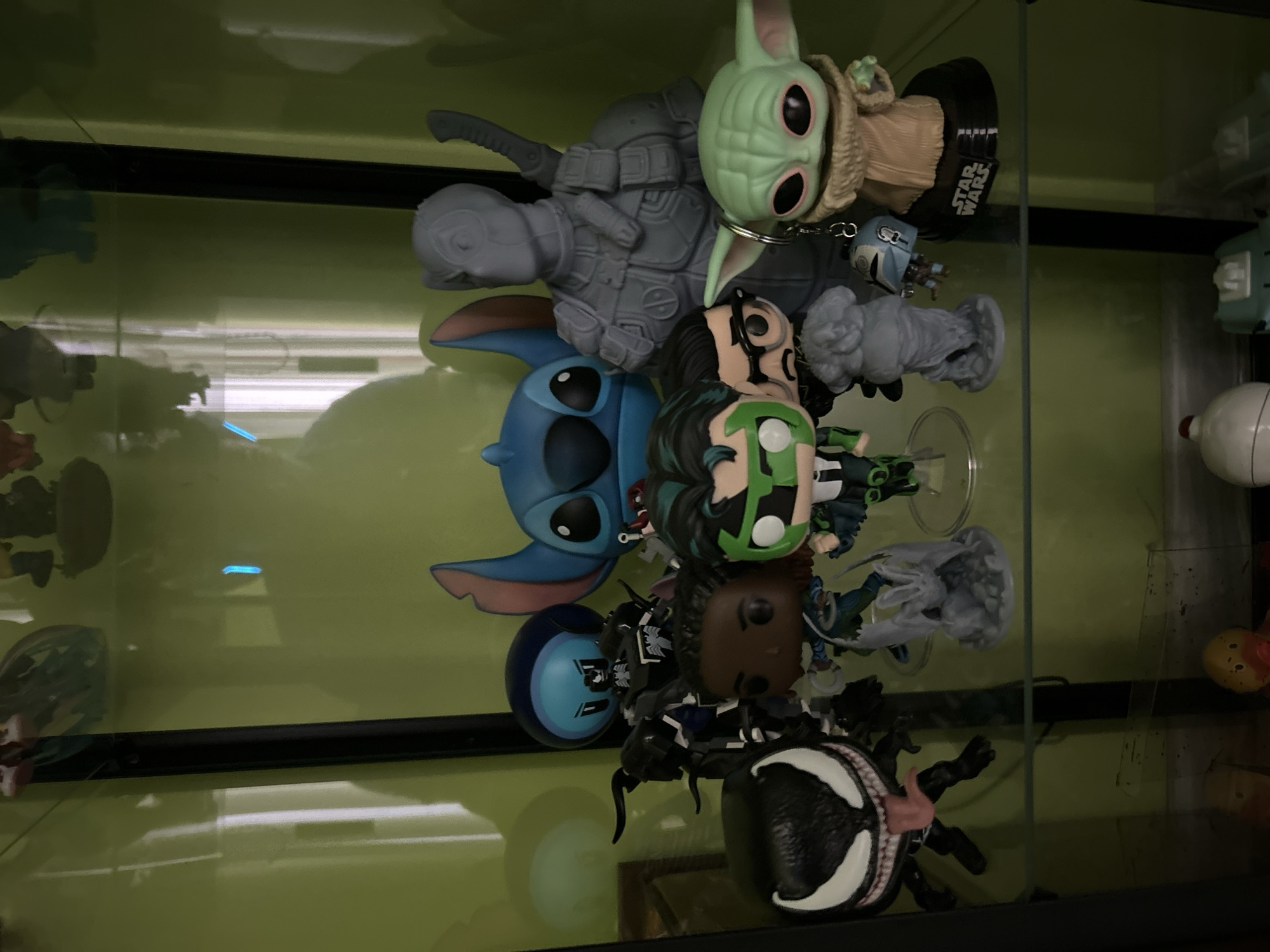 A collection of Funko Pop figurines is densely packed together on a glass shelf. The figures include "The Mandalorian," "Grogu," "Stitch," "Izuku Midoriya," "Venom," and several others that are difficult to identify due to the angle and proximity of the figures. The environment appears to be inside a glass display case, likely in a home or retail setting. The setting is not in Plovdiv, Bulgaria.