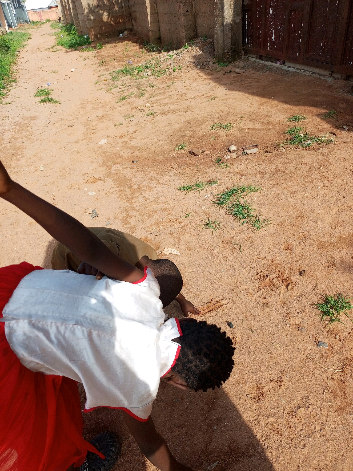 In Tempure, Nigeria, two children are engaged in an activity on a dirt path. One child, wearing a white top with red trim and a red skirt, is kneeling and appears to be drawing or writing in the dirt with their finger. Their dark, curly hair is visible. Another child, wearing a tan shirt, is also kneeling on the ground, facing away from the viewer. The surroundings consist of a sandy or dirt path, with sparse patches of green grass and small rocks. To the right of the path, there is a wooden gate or door with a textured surface, and a brick wall extends to the left. The scene is illuminated by sunlight, casting shadows on the ground.