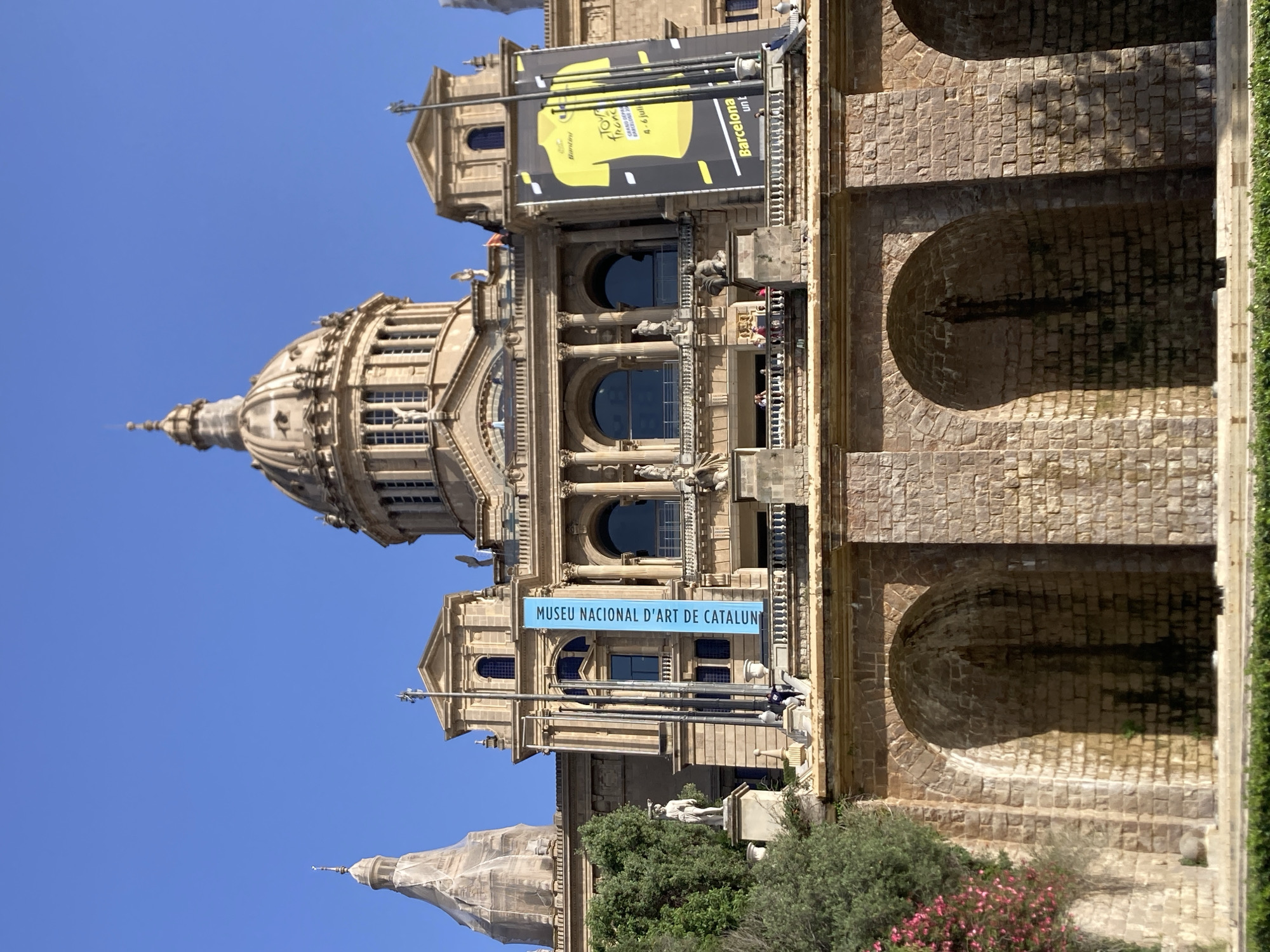The image captures the National Art Museum of Catalonia, located in Barcelona, Spain. The building is a grand, palatial structure with a large central dome and two smaller domes on either side. The facade is made of stone, with ornate architectural details, including columns, arches, and statues. In front of the building is a tiered stone terrace with people visible on it. There are also two large banners hanging from the building. One banner, on the left, is blue and has white text that reads "Museu Nacional d'Art de Catalunya". The other banner, on the right, is yellow and features an image of a yellow t-shirt with black and white graphics. Below the terrace, there is a stone wall with large arched alcoves. The sky is a clear, bright blue, indicating a sunny day. Lush green trees and pink flowering bushes are visible on the left side of the image.