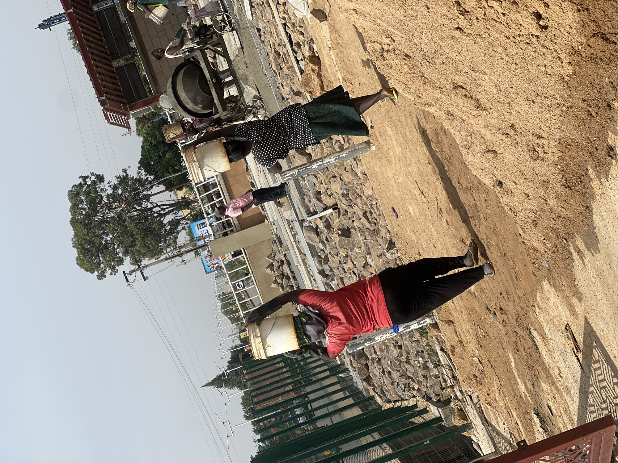 In Barakin Lerawi, Nigeria, two women carry buckets on their heads near a construction site. One woman wears a red shirt and black pants. The other wears a black and white polka dot shirt and a green skirt. A pile of dirt is in the foreground. A concrete mixer and other construction equipment are visible in the background, along with buildings, trees, and utility poles.