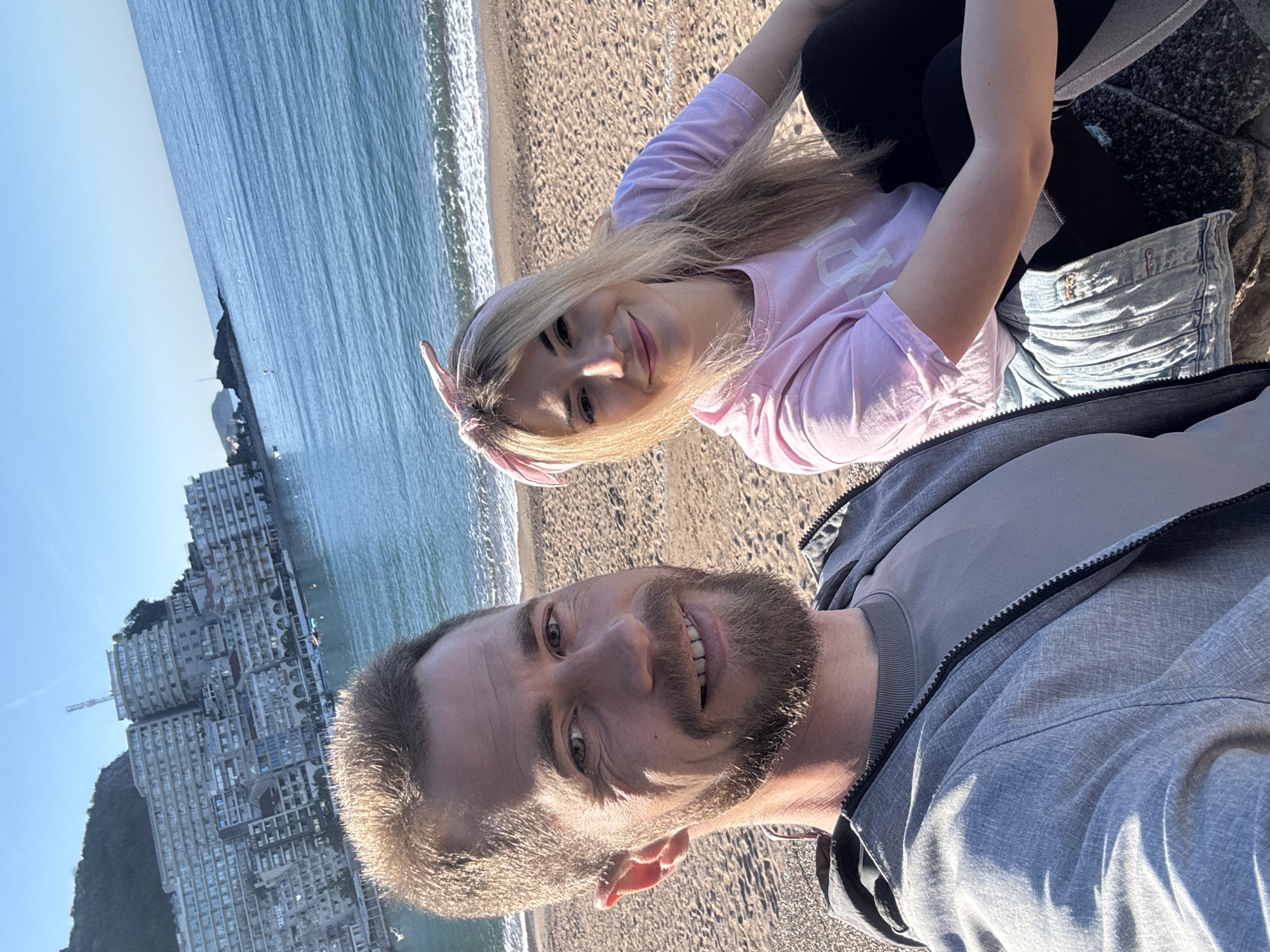 A selfie shows a man and a woman at a beach in Rafailovići, Montenegro. The man, in the foreground, is smiling and wearing a gray jacket and gray t-shirt. The woman, behind him, is also smiling and wearing a pink t-shirt and jean shorts, with a pink headband. They are likely sitting or kneeling on a rocky area next to the sandy beach. The Adriatic Sea and a building complex are visible in the background. The photo is taken in bright daylight.