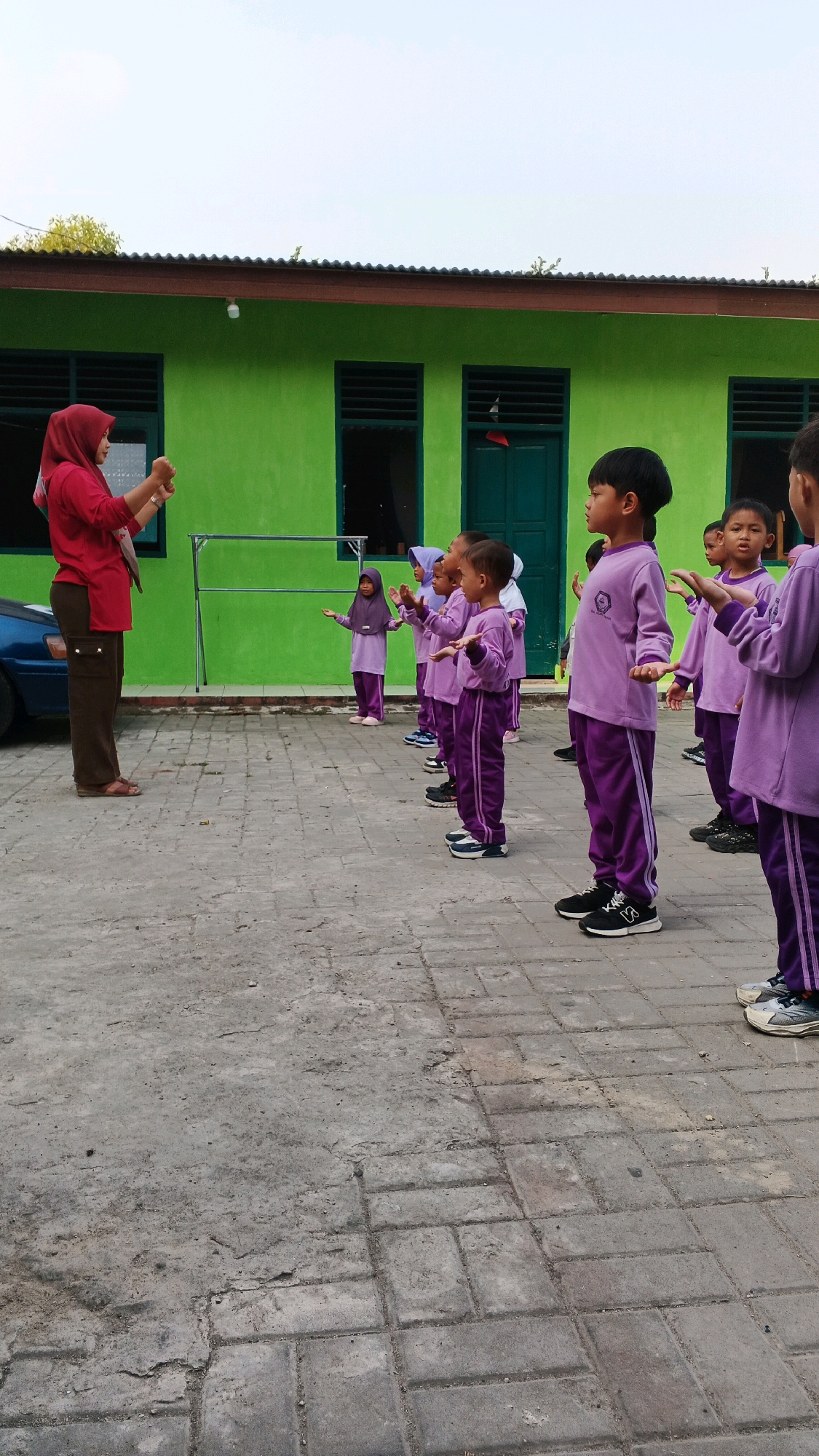 In front of a bright green school building with dark brown trim and dark green doors and windows, a woman in a red hijab and a red shirt stands facing a group of children in purple uniforms. The children, who appear to be preschoolers, are lined up, facing the woman. The woman is holding her hands up as if demonstrating an action. The children are also holding their arms out. The ground is paved with grey interlocking bricks. A blue car is partially visible on the left side of the image. The image was taken in the city of Serang, Indonesia.