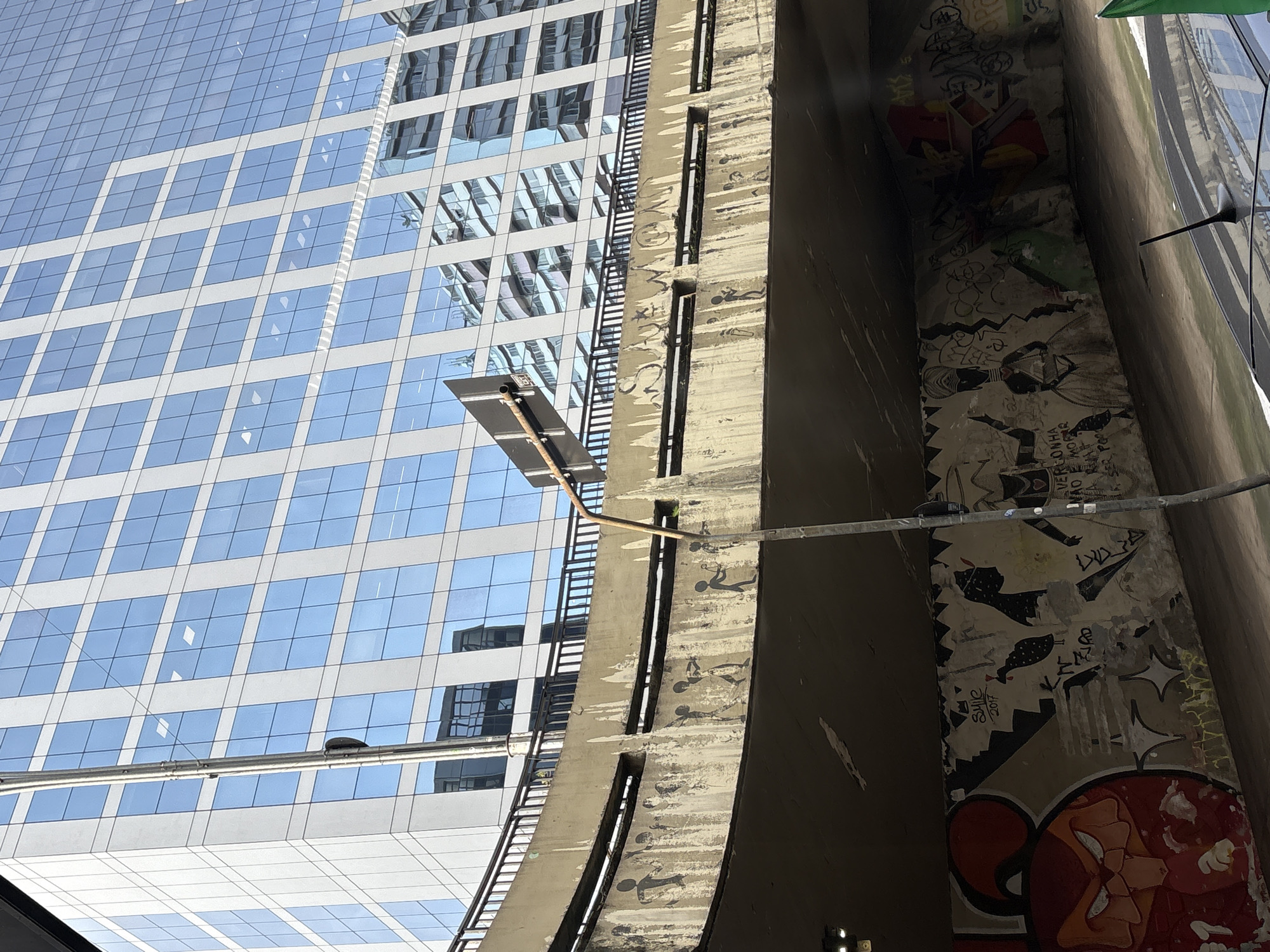 The image shows a street-level view looking up at an elevated highway overpass and a modern glass-fronted skyscraper in São Paulo, Brazil. The overpass is concrete and adorned with graffiti, featuring stylized figures and text. Below the overpass, the wall is also covered in extensive graffiti, including a large red cartoon-like character on the left and various drawings and tags. A light pole extends upwards on the right side of the frame. The skyscraper in the background has a grid-like facade of blue glass windows. Reflections of other buildings are visible in the skyscraper's glass. The scene appears to be in an urban environment, likely a busy thoroughfare given the presence of the highway and the scale of the buildings.