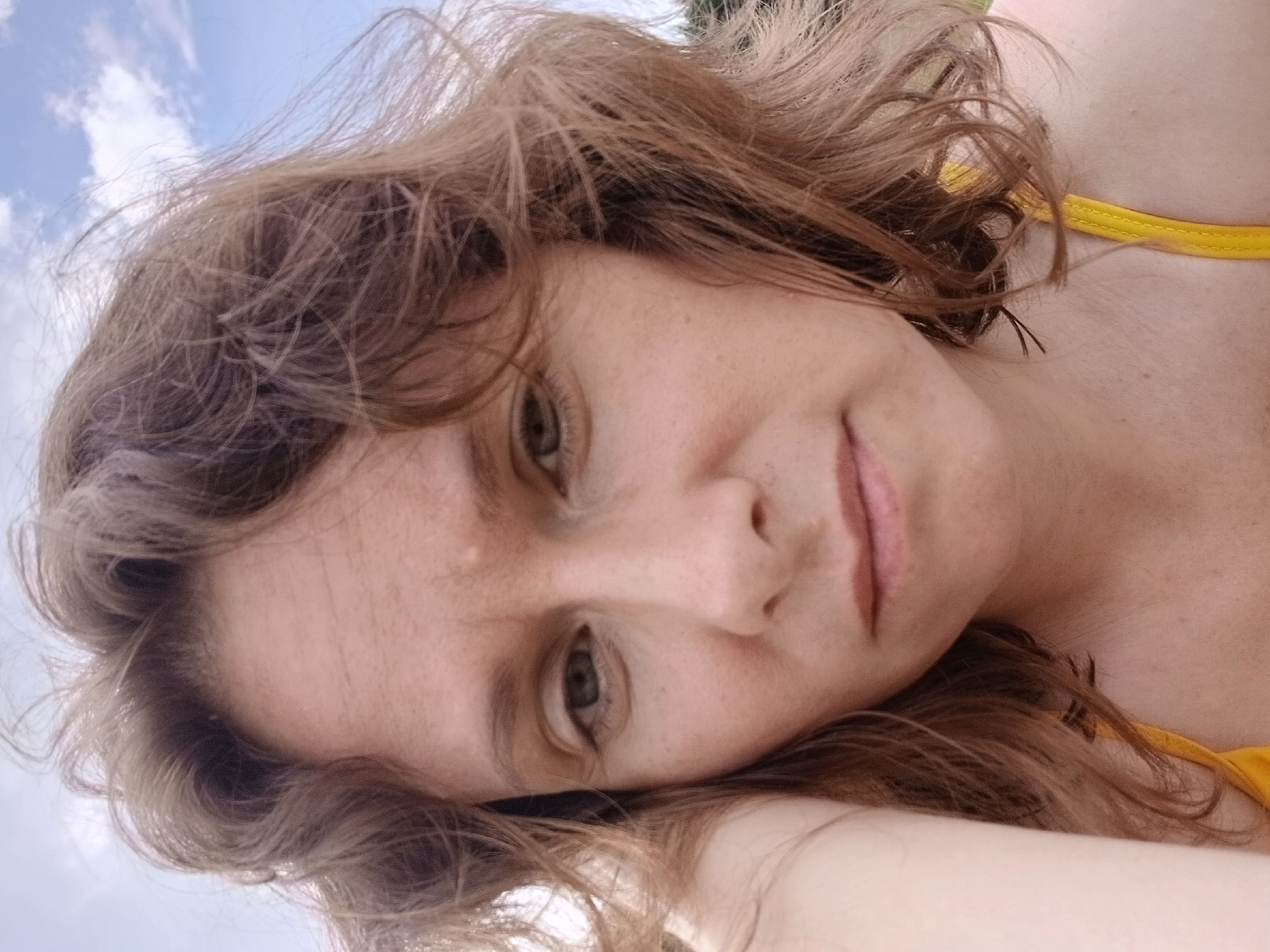 The image shows a close-up, selfie-style shot of a woman with light brown, wavy hair. She has a fair complexion with freckles and is looking slightly off-camera with a neutral expression. Her left arm is visible resting near her face. She is wearing a yellow top. The background appears to be outdoors with a cloudy sky. There is no discernible urban environment or specific location information provided to indicate a connection to Revda, Russia.
