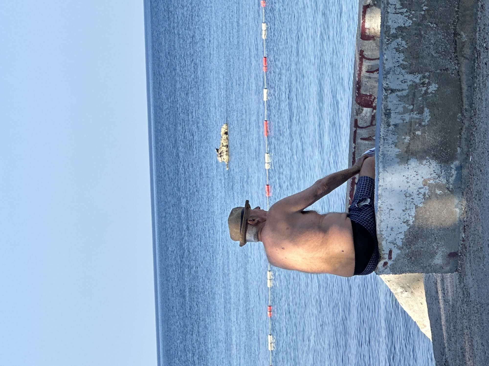 An elderly man wearing a straw hat and checkered swim trunks sits on a concrete barrier facing a calm, blue sea. He is shirtless, revealing his sun-tanned back. In the background, a buoy line with alternating red and white floats stretches across the water. Further out, a small rocky outcrop is visible. The scene is set on a bright, clear day. The man is not interacting with anyone and his action is simply sitting and looking out at the sea.