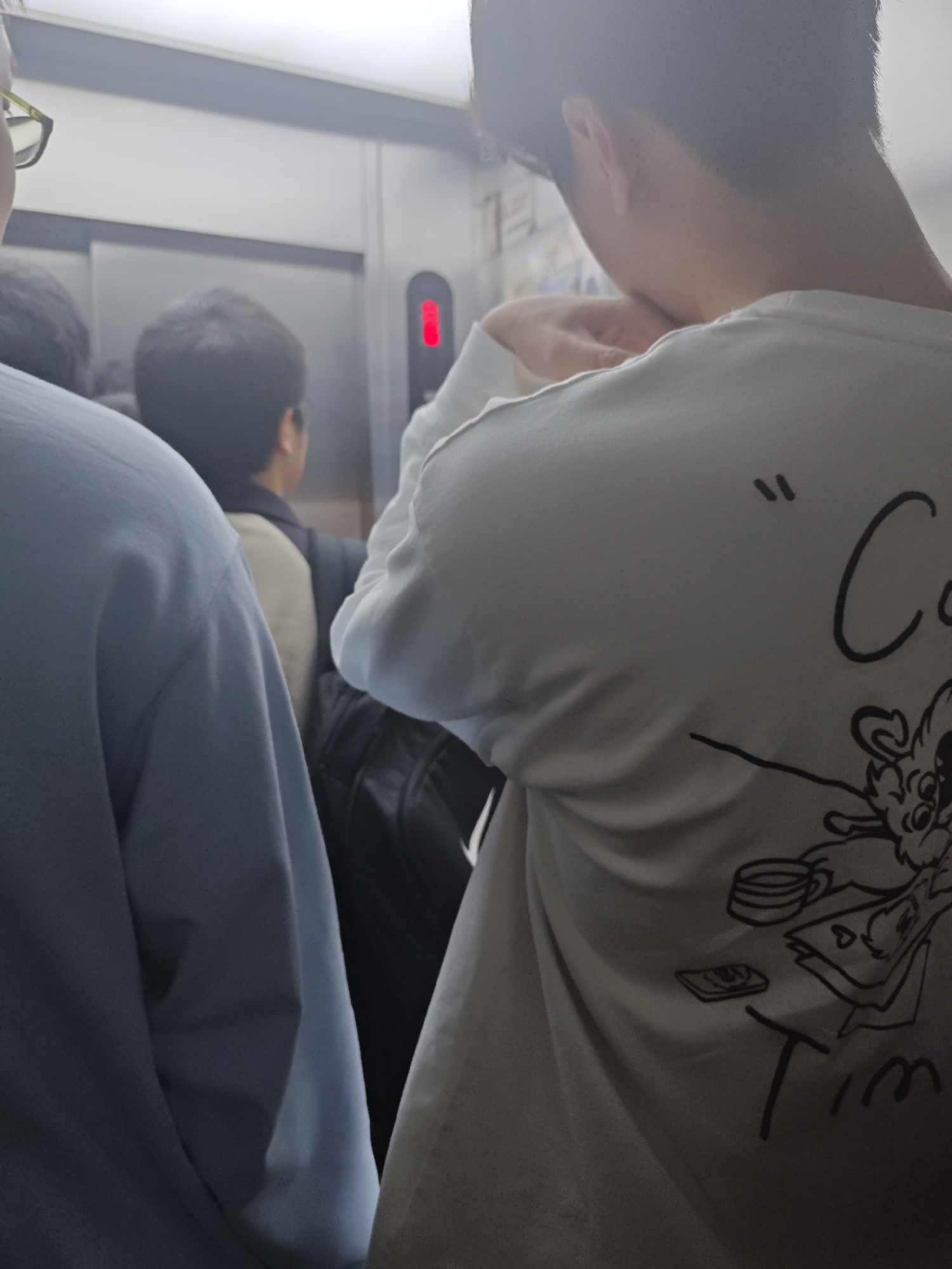 The image captures the crowded interior of an elevator in Yuzhong District, China. In the immediate foreground, a person is seen from the upper back, wearing a white long-sleeved shirt adorned with a black line art graphic. The graphic depicts a stylized creature, a coffee cup, stacked papers with a heart symbol, a small rectangular object, and the word "Time." This individual's left hand is raised to their neck/collar area. To the left, the shoulder and part of the head of another person are visible, wearing a light blue long-sleeved garment and green-rimmed eyeglasses. Between these two, a third person is observed from the rear, wearing a dark jacket over a light-colored top and carrying a dark backpack. The elevator's metallic gray doors are closed, and a control panel on the right side displays a red illuminated digit. The overhead lighting creates a bright but somewhat diffused illumination within the confined space.