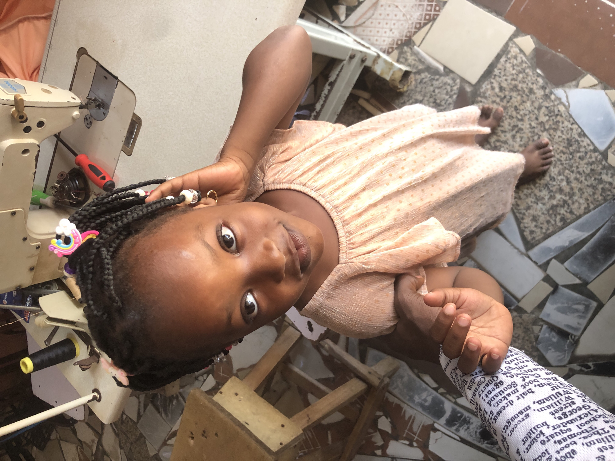 A young girl with dark skin and braided hair stands in front of a sewing machine in a workshop in Ado, Nigeria. She wears a peach-colored dress and has colorful hair accessories. The girl is looking directly at the camera with a slight smile, her right hand touching her ear. Beside her, a hand wearing a white sleeve with black text is reaching out. The background shows a sewing machine, a spool of black thread, a red screwdriver, and a wooden stool. The floor is tiled with a mosaic of different colored stones and tiles.