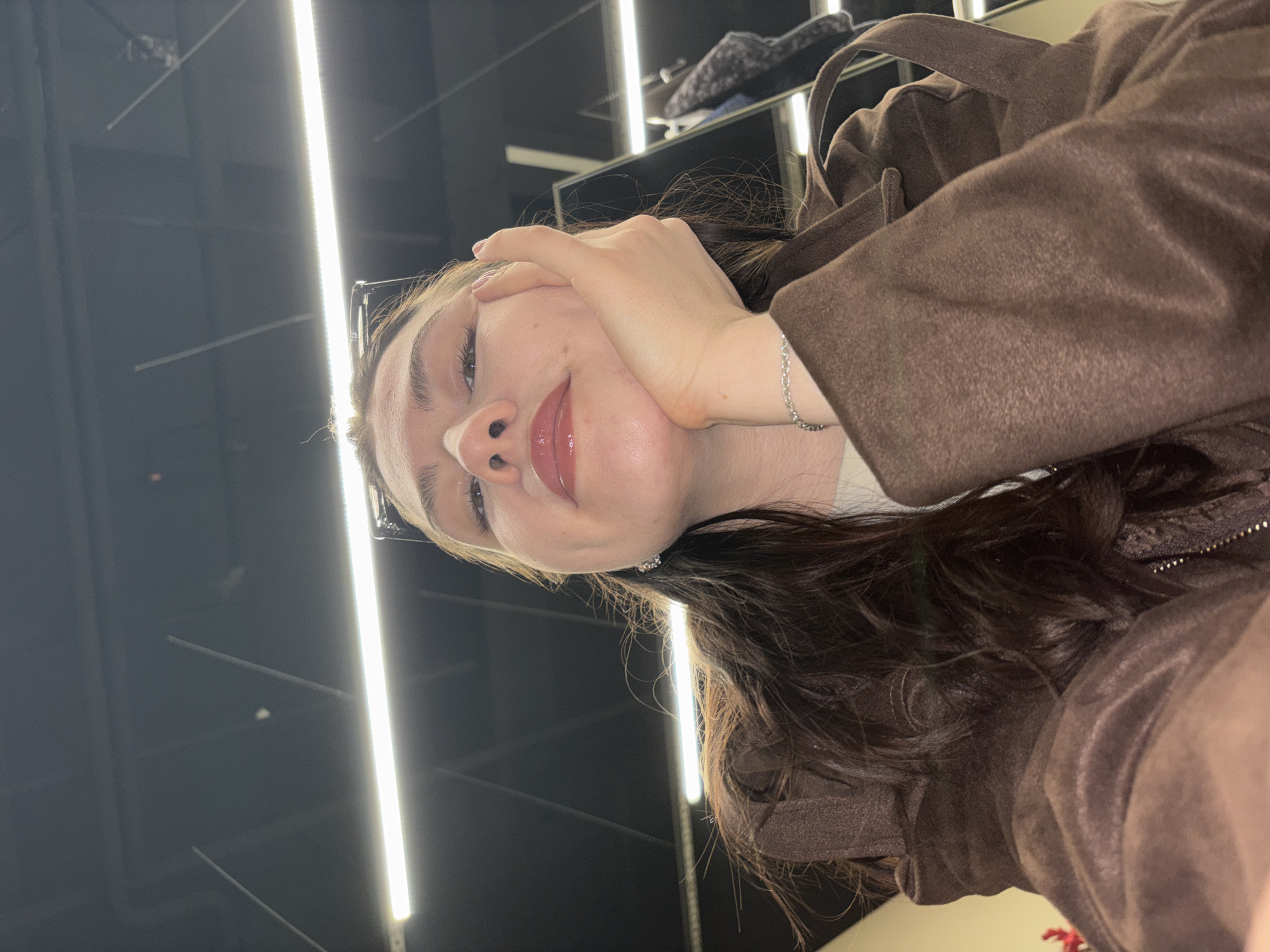 An upward-angled, close-up shot captures a young woman from the chest up in an indoor setting, likely in Perm, Russia. She has long, dark hair styled in waves and fair skin. Her light-colored eyes are looking slightly upwards, and a subtle smile plays on her lips, accentuated by a clear lip gloss. A pair of clear-framed glasses is pushed up onto her head. Her left hand is gently placed against her cheek, with her fingers resting near her temple. A delicate silver chain bracelet is visible on her left wrist.
She is dressed in a brown jacket, which appears to have a soft, suede-like texture. The background is predominantly dark, featuring several distinct, bright vertical linear light fixtures, characteristic of modern commercial or public spaces like a store or exhibition hall. These lights provide the primary illumination, creating a contrast with the otherwise dim surroundings. Faint, dark forms in the background suggest shelving or structural elements. The overall impression is one of casualness and calm. No visible text is present in the image.