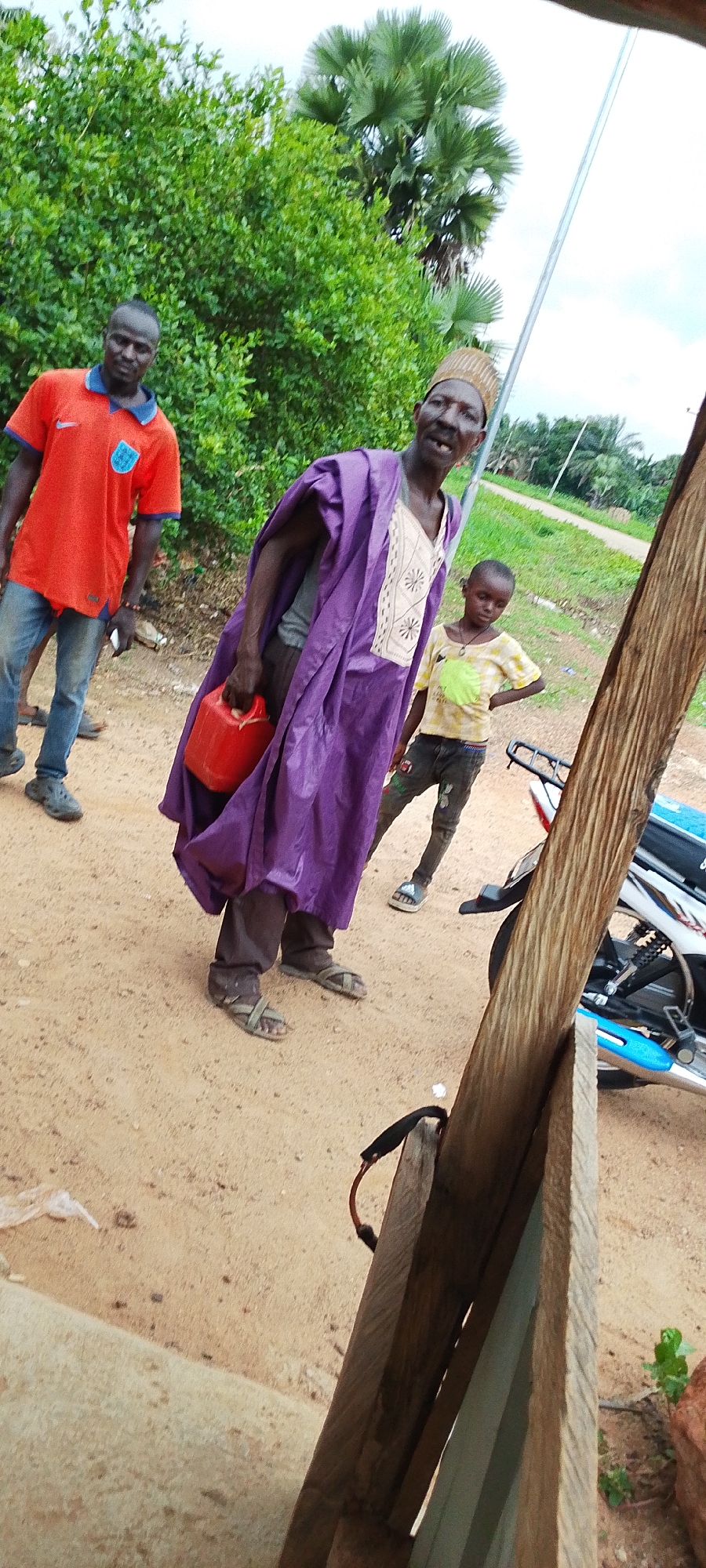 The image depicts an outdoor scene in Kamantan, Nigeria. In the foreground, a wooden structure partially obstructs the view. Two men and a child are visible. The man on the left wears an orange t-shirt and blue jeans, with dark skin and hair. He is looking towards the camera. The central figure, an older man, is dressed in a vibrant purple robe with a patterned neckline and a straw hat. He holds a red jerrycan in his left hand and looks at the camera with an open mouth. To the right of the central figure is a young child wearing a yellow and green tie-dye t-shirt and dark pants, with their hands on their hips. Behind them, lush green trees and palm trees form the backdrop. A motorcycle is partially visible to the right of the central figures. The ground is sandy and appears to be a dirt road. The overall lighting suggests an overcast day.