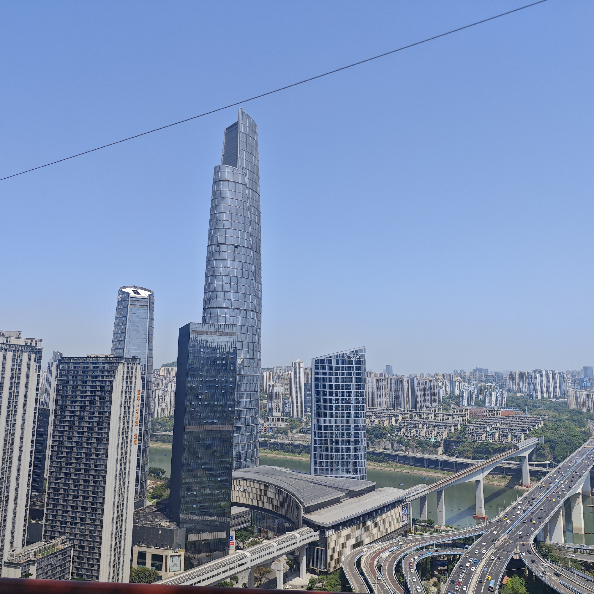 An elevated wide shot shows the cityscape of Yuzhong District, Chongqing, China, with modern buildings, a river, a bridge, and clear blue sky. The most prominent building is a tall, glass-paneled skyscraper in the center of the image. Several other skyscrapers and tall buildings surround it. The Yangtze River flows horizontally in the midground. The bridge has multiple lanes of traffic and is supported by concrete pillars. A network of roads and on-ramps with cars is visible below the bridge. The background shows a densely populated urban area with many residential buildings. The sky is clear and blue with no clouds. A dark wire stretches across the top left portion of the image.