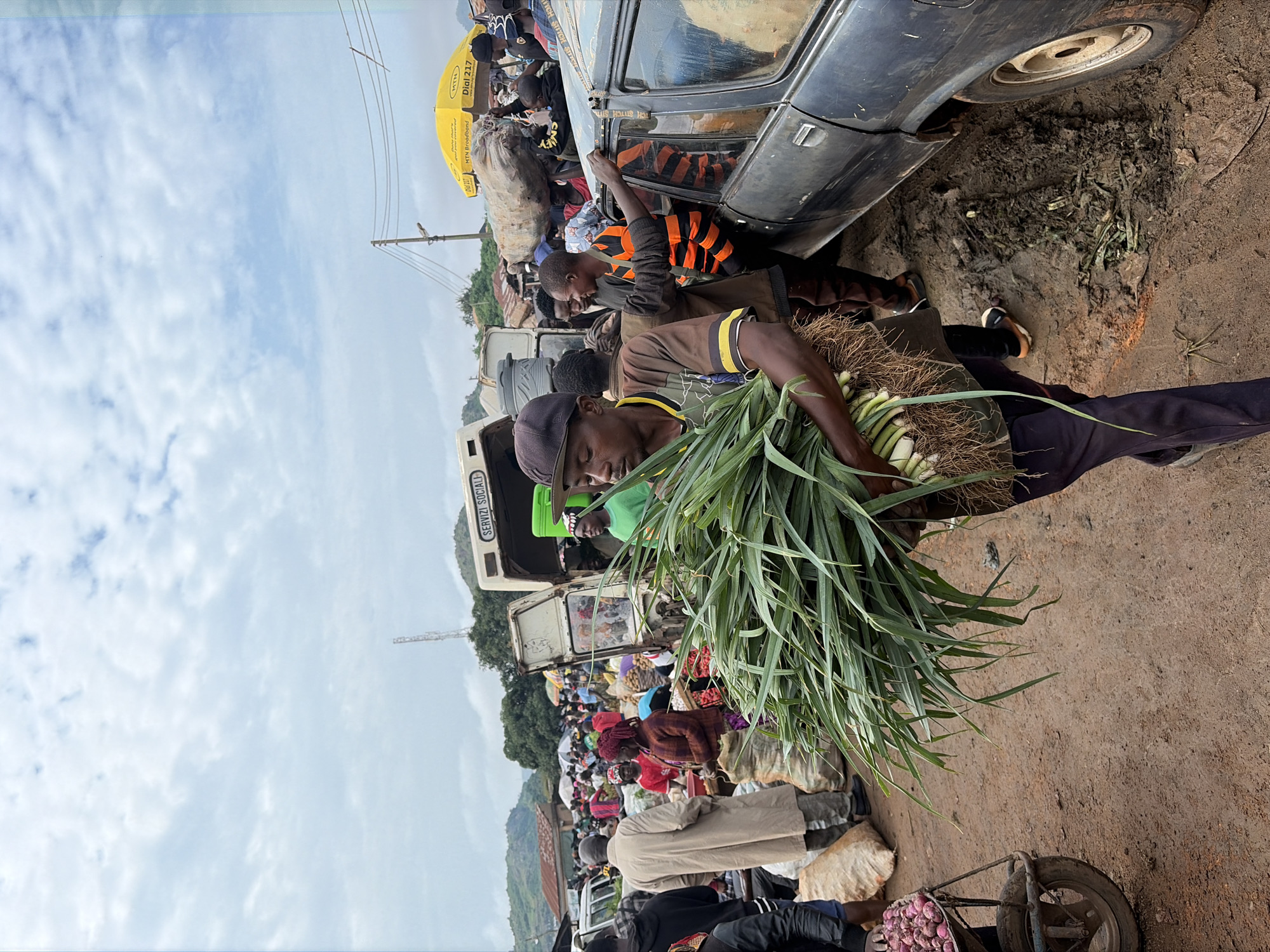 In Gura Top, Nigeria, a bustling outdoor market is underway. The sky is overcast with scattered clouds. In the foreground, a man is carrying a large bunch of green leafy vegetables. He is wearing a brown and yellow t-shirt and dark pants, with a cap on his head. To his right, a dark-colored car is parked on a muddy patch of ground. A man in an orange and black striped shirt is leaning against the car. Behind them, a busy market scene unfolds with numerous people, stalls, and vehicles. A white van with "SERVIZI SOCIALI" written on it is parked in the background. A yellow umbrella with "MTN" branding is visible further back. The ground is dirt and appears wet and muddy in places, suggesting recent rain. Various goods, including sacks of produce and onions in a wheelbarrow, are visible throughout the market. The overall atmosphere is one of lively commerce and daily life in a Nigerian market.