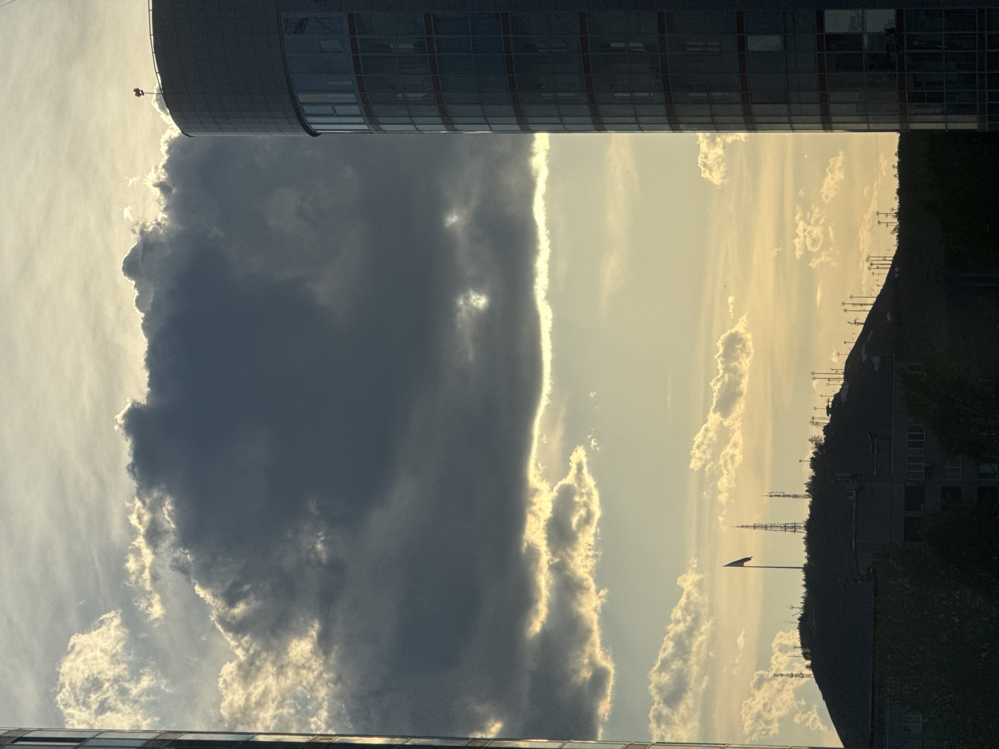 The photograph captures a view in Krasnoyarsk, Russia, framed by two buildings, one possibly cylindrical and glass-covered. A dark, imposing cloud formation fills much of the left side, contrasting with the bright, sunlit sky on the right. The skyline includes a wooded hill with communication towers and a Russian flag, set against a backdrop of light clouds.