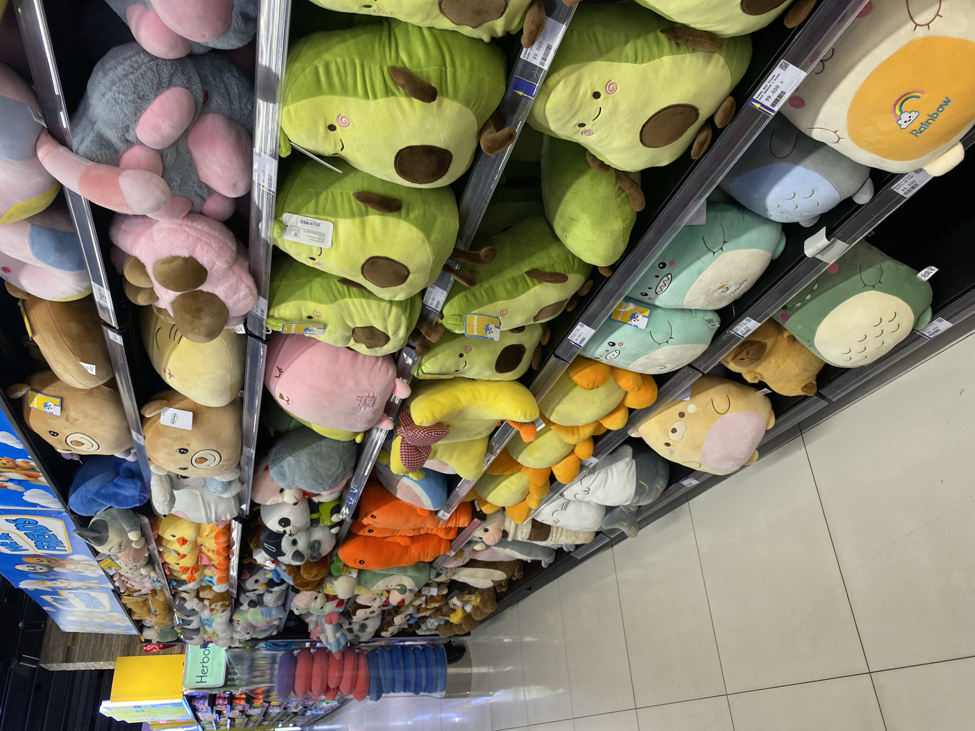 The image shows a brightly lit aisle in a retail store, likely a department store or a supermarket with a toy section, displaying a wide variety of plush toys on tiered shelves. The toys are predominantly large and colorful, featuring cartoonish designs such as avocados, bears, ducks, and other animals. Some toys are stacked, while others are arranged individually. The shelves are made of dark material, possibly metal, and are well-stocked. In the background, a large sign with cartoon characters and text is visible, suggesting a themed display. There are no people visible in the image. The flooring is a light-colored tile. The general ambiance is one of a commercial retail environment designed to attract shoppers, particularly children. The provided information about the location in Banda Aceh, Indonesia, does not appear to be visually verifiable within the image itself.
