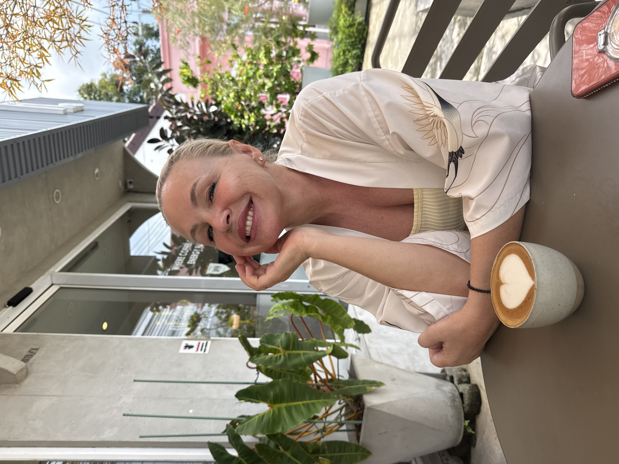 A blonde woman sits at a table outside a cafe in Koh Samui District, Thailand. She is smiling at the camera and resting her chin on her hand. The woman is wearing a light-colored kimono with a bird pattern and a yellow top underneath. In front of her is a cup of coffee with latte art in the shape of a heart. The background shows a cafe entrance with glass doors, a large potted plant, and some greenery. The cafe appears to be on a street, with a pink building visible in the distance. A red mobile phone is placed on the table next to the coffee cup.