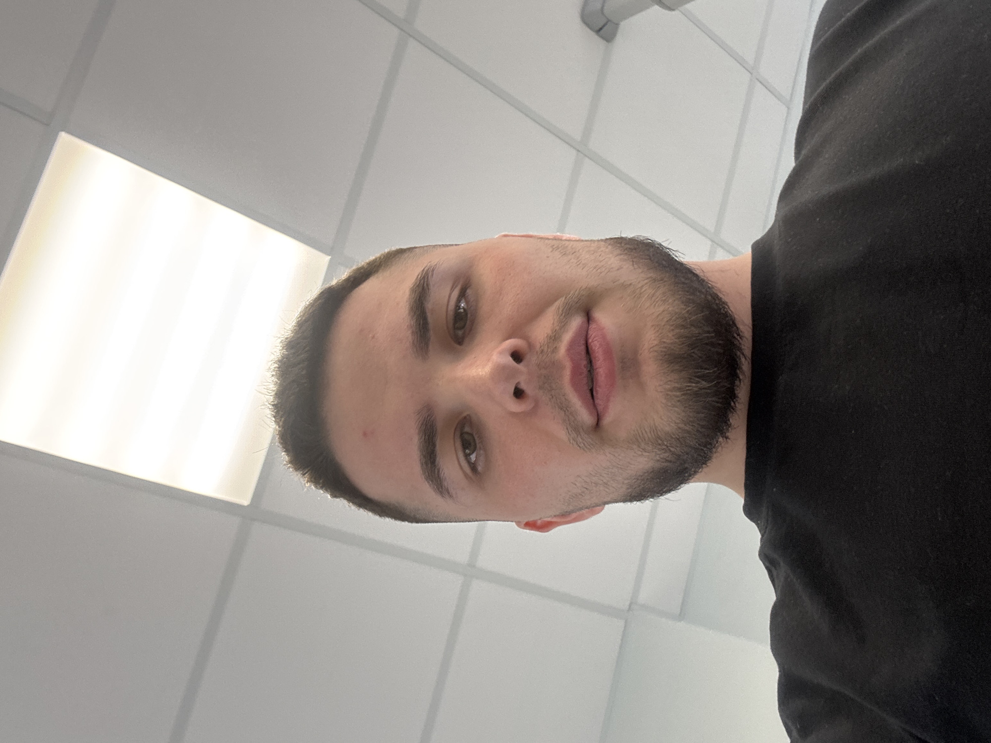 The image captures an upward-angled selfie of a young man indoors in Perm, Russia. He has short dark hair, a neatly trimmed beard or stubble, and brown eyes, looking directly at the camera with a neutral expression that hints at a subtle smile. He is wearing a plain black t-shirt. The background features a grid-patterned dropped ceiling with light-colored, possibly white or light grey, tiles, typical of an office, school, or commercial building. A large, bright rectangular light fixture, likely a fluorescent panel, is visible in the upper part of the frame, providing ample artificial illumination for the scene. No specific time of day or outdoor weather conditions can be determined from the indoor setting. There is no visible text.