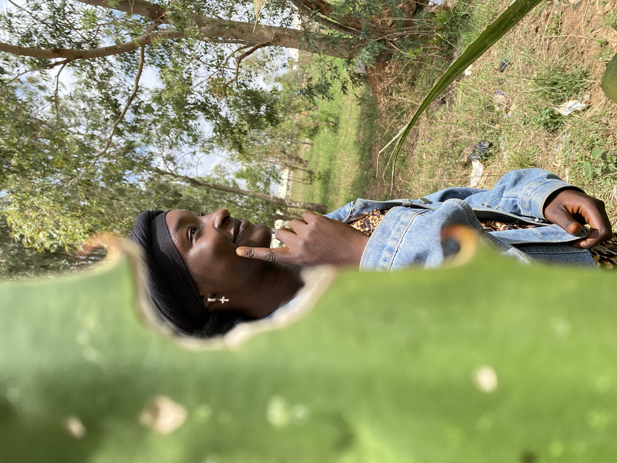 A young person with dark skin is lying on their side in a grassy outdoor environment in the city of Durimi, Nigeria. They are wearing a black head covering, a denim jacket, and a patterned top. A silver cross earring is visible. Their left hand is near their face, and they are looking upwards. The foreground is blurred by a large green plant leaf, possibly an aloe vera plant. The background consists of trees, grass, and vegetation, with some litter scattered around. The sky is visible through the trees.