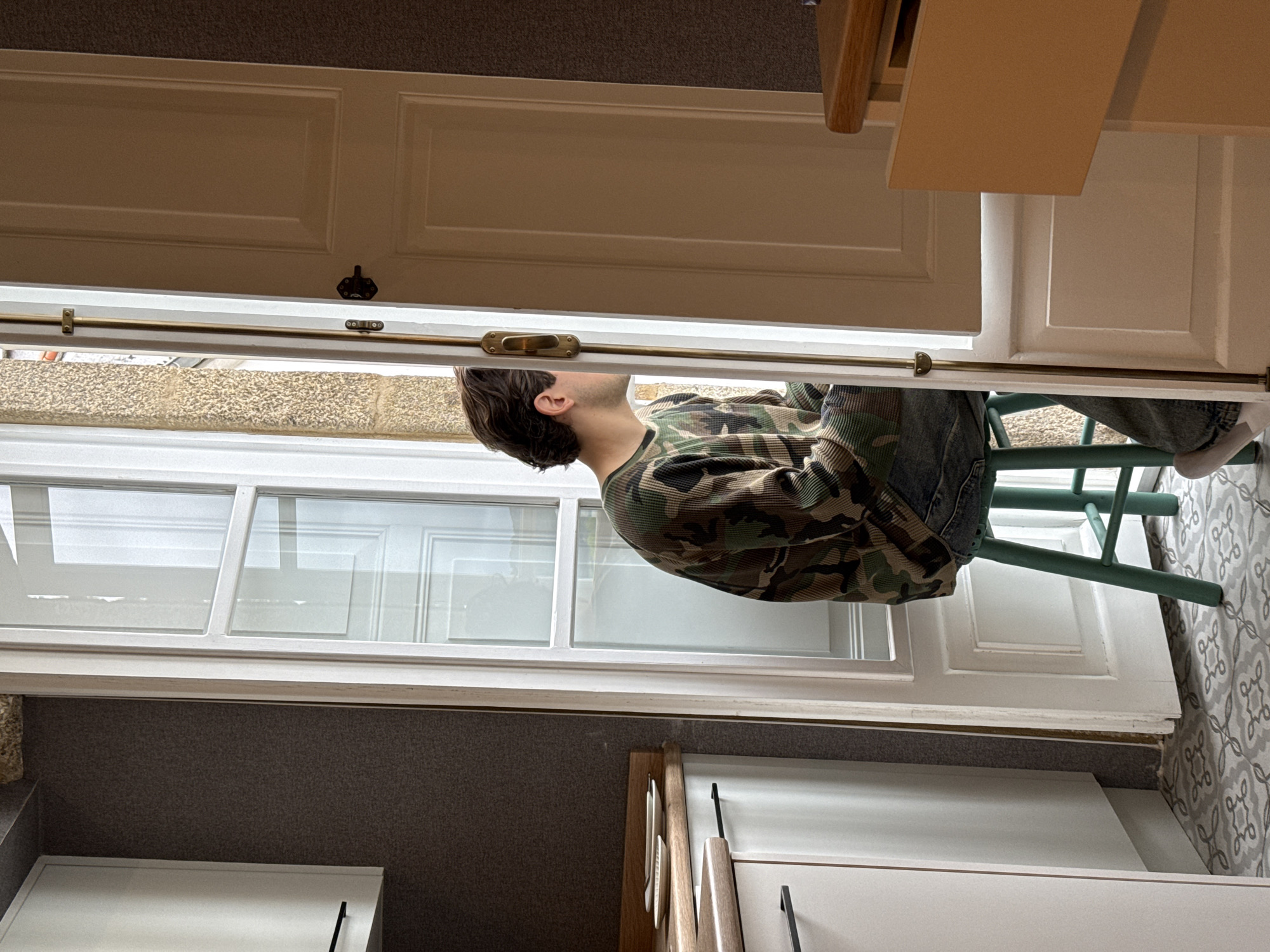 The image depicts a person sitting on a green chair and looking through an open door in what appears to be an indoor setting. The person is wearing a camouflage long-sleeved shirt and jeans, with fair skin and dark hair. The door opens to an exterior view of a multi-paned window, and above the window, a wall of light-colored stone is visible. The foreground shows the inside of a room, with white cabinets and gray flooring, while the background reveals a patterned tile floor. It is unknown if this photo was taken in Santiago De Compostela, Spain.