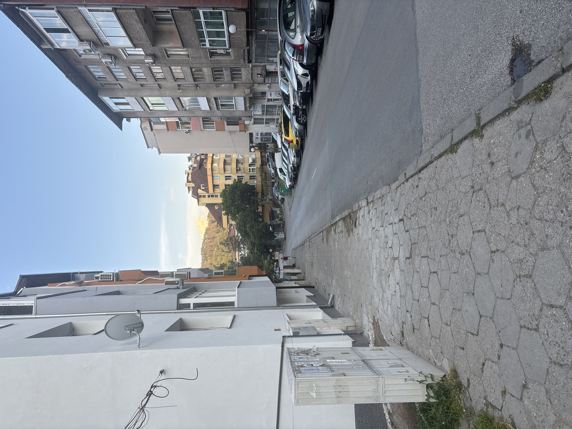 The image displays an urban street scene in Blagoevgrad, Bulgaria, under a clear blue sky during daylight hours. The perspective is looking down a street bordered by multi-story residential buildings.
On the immediate left, a modern, light-colored building is prominent, featuring white and grey sections, a visible satellite dish, and several air conditioning units attached to its facade. Beyond this, further along the left side of the street, stand older, grey concrete apartment blocks characterized by numerous windows, balconies, and additional external air conditioning units.
The street itself is an asphalt road with a continuous line of parked cars on the left side, including various sedans and hatchbacks such as a grey Peugeot 307 and a distinct yellow car. The license plates are in a European format, consistent with the specified location.
On the right side of the street, the sidewalk is paved with a combination of hexagonal concrete blocks and rectangular pavers, showing signs of age and wear, with weeds growing in crevices. In the background, past the buildings and trees, a large, forested hill or mountain rises under the clear sky, providing a natural backdrop to the urban setting. Buildings on the right side of the street also appear to be residential, some with facades in shades of yellow and brown.
No specific activity is clearly depicted, aside from the parked cars and a very small, indistinct figure walking in the distance on the right sidewalk in one of the hint crops. The scene portrays a typical residential neighborhood with a mix of architectural styles. No legible text is visible on signs or vehicle license plates in the main image.