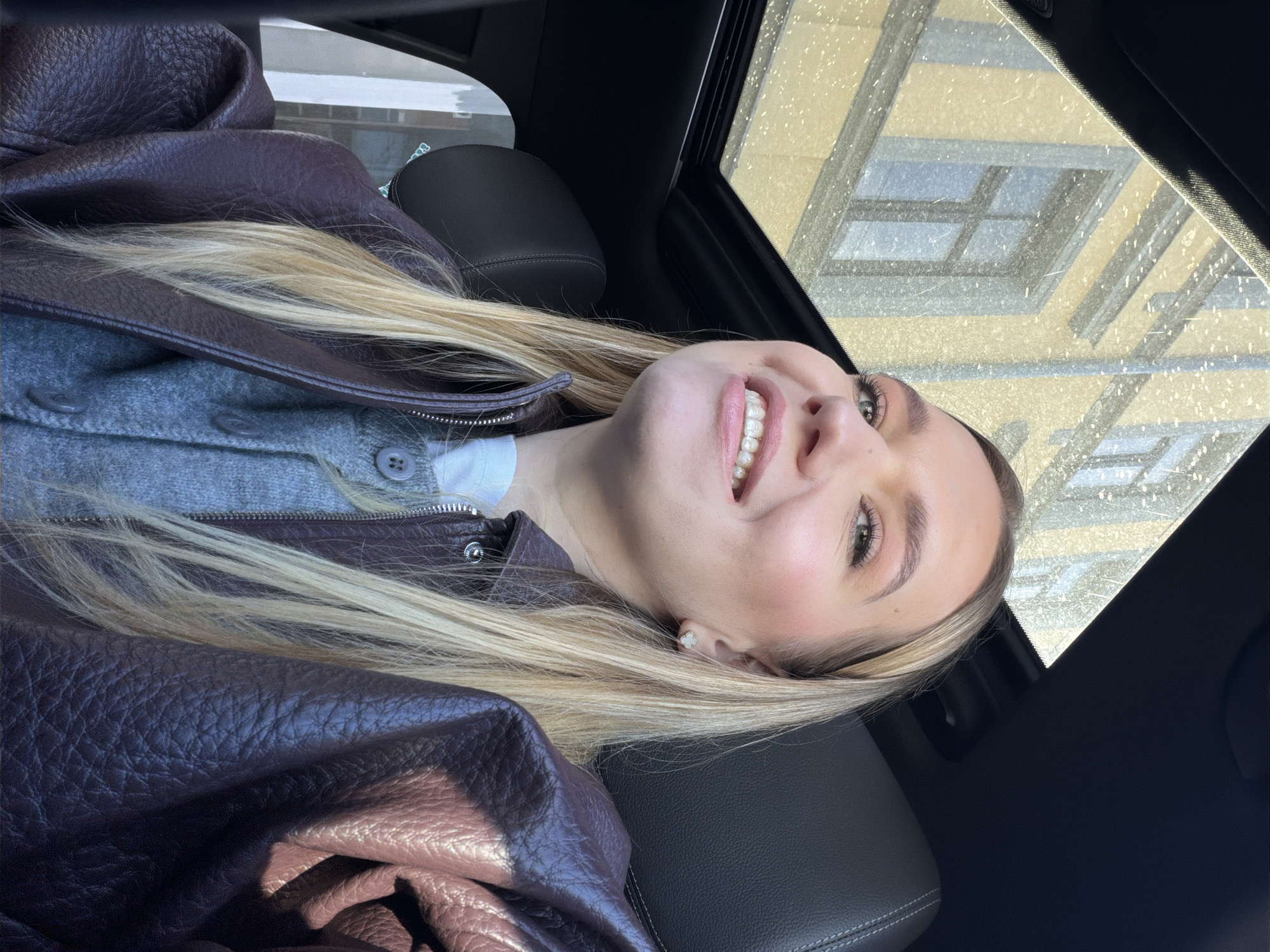 A fair-skinned young woman with blonde hair is lying in a car, looking upwards with a smile. She is wearing a brown leather jacket over a grey cardigan and a white shirt. The car's sunroof is open, revealing a building exterior with windows, likely in Riga, Latvia, given the architectural style. The interior of the car features black leather seats. The image has a slightly rotated orientation. Raindrops are visible on the sunroof.