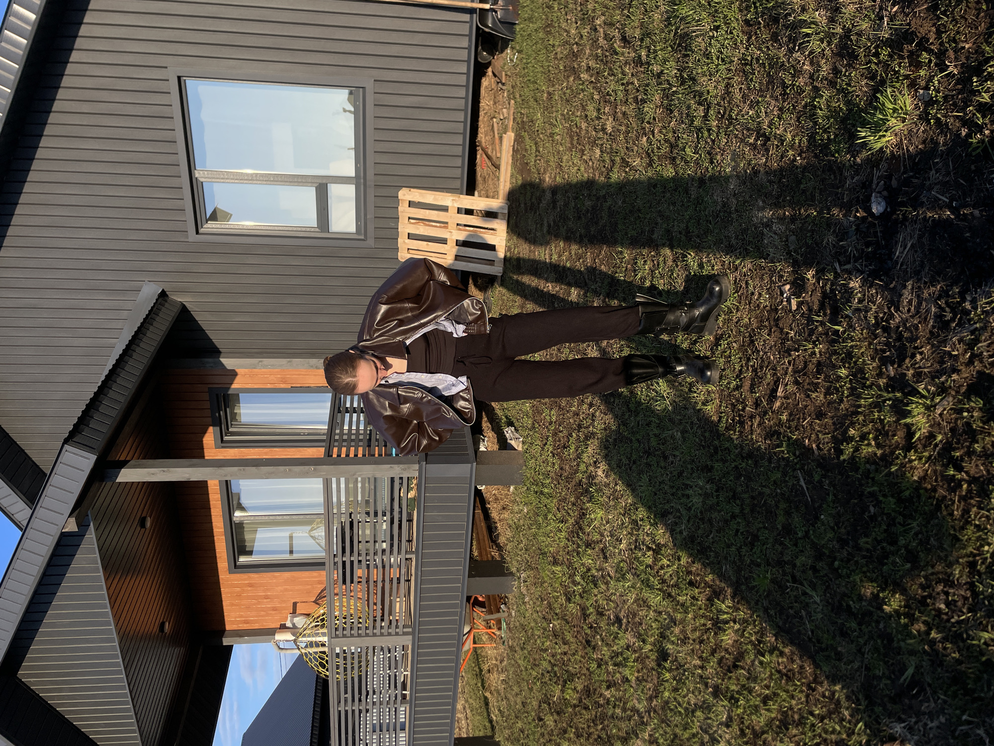 An outdoor, rotated image depicts a person standing near a modern house in what appears to be a residential or semi-rural setting in Podshivalovo, Russia. The person, positioned centrally, is wearing a brown leather jacket, dark trousers, and dark boots. Their head is tilted, and their face is partially obscured by shadow.
The house behind them features a contemporary design with dark grey horizontal siding on the upper level and dark brown, wood-textured siding below. Multiple windows with dark frames reflect a clear, bright sky. A raised balcony or deck with dark grey vertical railings is visible on the lower level, and a yellow spherical object (possibly a hanging chair or decoration) is partially seen on it.
The ground is covered with patchy grass and bare earth. Long, distinct shadows are cast across the grass, indicating either early morning or late afternoon sunlight. A wooden pallet rests on the ground near the house, with other construction-related materials partially visible in the background. The weather appears sunny with clear skies. There is no visible text in the image.