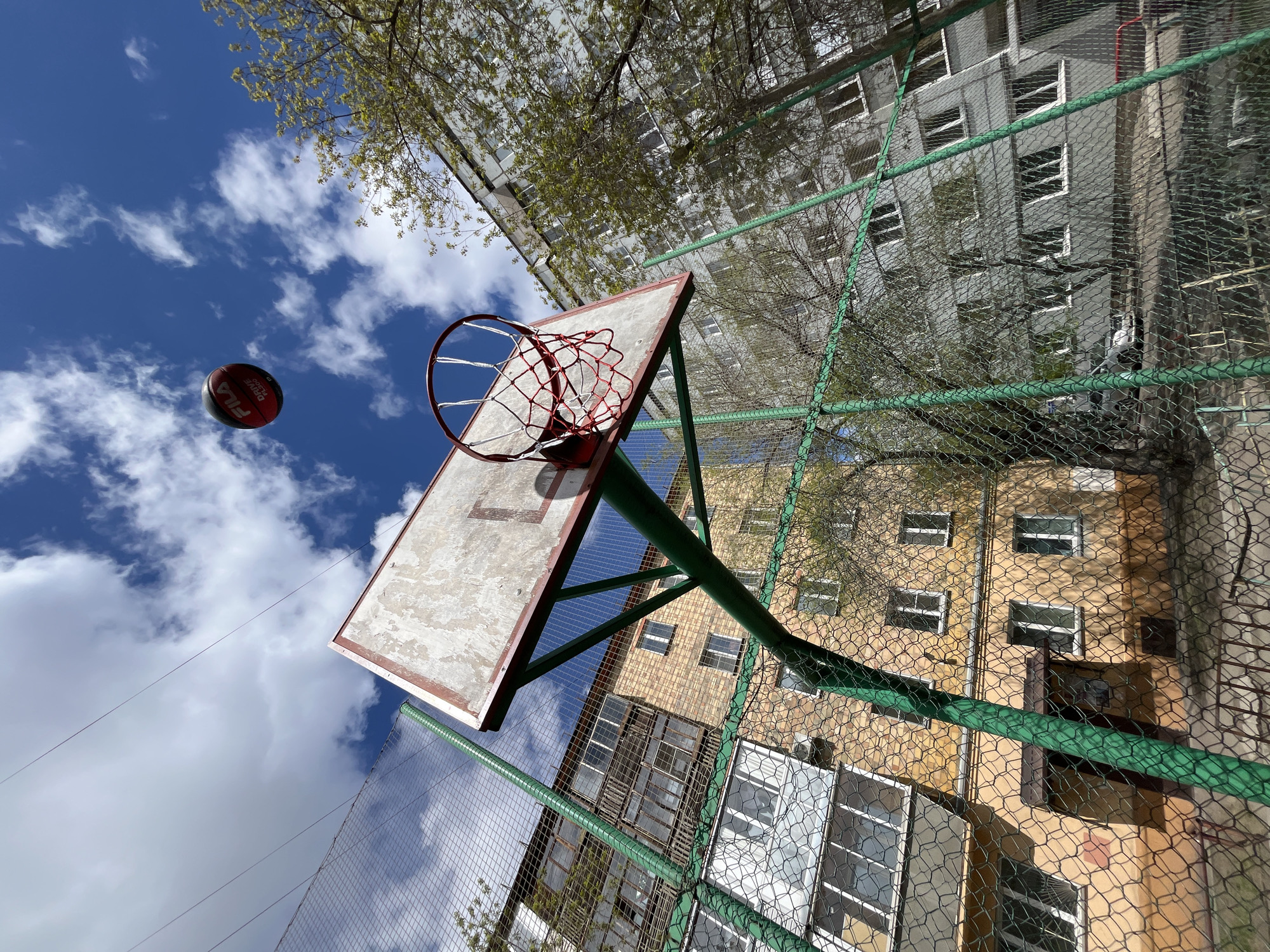 The image shows a basketball hoop with a red and white net, against a backdrop of two apartment buildings in Krasnoyarsk, Russia. The building to the left is yellow, with multiple windows and balconies. The building behind it is grey with similar window placement. A green chain-link fence surrounds the basketball court. A red and black basketball, a "Fila Drive Performance" is mid-air to the left of the hoop, under a partly cloudy blue sky. Bare tree branches are visible near the top of the buildings. The photo appears to be taken from below, angling upwards.
