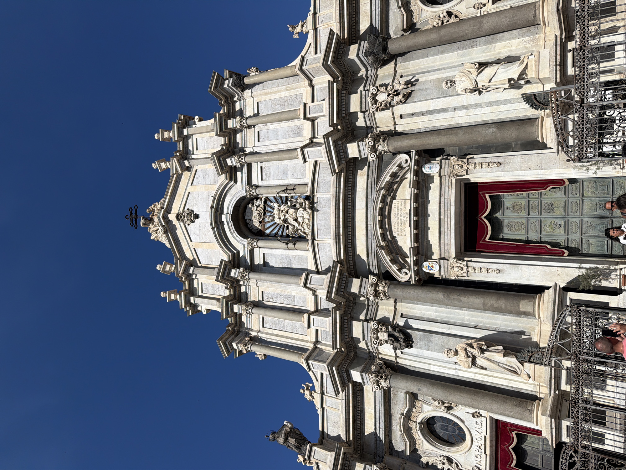This is a low-angle, full-frame, eye-level shot of the front facade of the Cattedrale di Sant'Agata in Catania, Italy, under a clear blue sky. The baroque-style cathedral is constructed of light-colored stone and features intricate sculptures, columns, and ornate details. A large, dark green double door with decorative metalwork is central to the image, flanked by red velvet curtains. Statues of figures in robes stand in niches on either side of the entrance. A man in a pink shirt is visible in the foreground, holding up a phone, seemingly taking a photo. Another person, partially obscured, is also visible near him. The cathedral's architecture is grand and imposing, with multiple levels and decorative elements visible. The scene appears to be a tourist location, with the presence of people and the focus on the historical building.