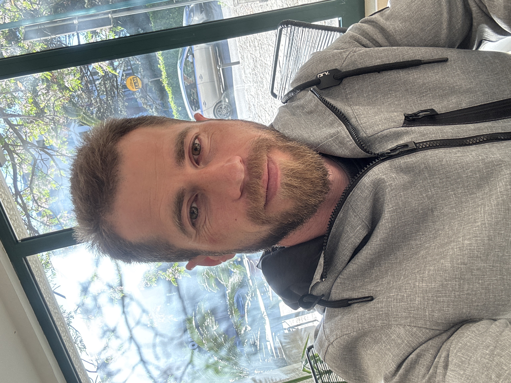 A man in his 30s or 40s is captured in a close-up, portrait-style shot, looking directly at the camera. He has short light brown hair, a short beard, and brown eyes, with a neutral to slightly smiling expression. He is wearing a grey, marled-patterned zippered jacket. A black zipper pull on his right shoulder bears the text "DRV."
The man is positioned indoors, in front of a large window that reveals an outdoor scene, indicating bright daytime with sunny weather. Through the window, green trees and foliage are visible, along with parts of an urban or suburban setting, including paved ground or a street. A white car and a light blue/grey van are parked outside. Reflections on the window's surface show a yellow circular sticker or sign with the text "L&M" and glimpses of the room's interior. A dark green or black window frame is prominent, running horizontally across the upper portion of the view. Part of a black metal outdoor-style chair is visible to the man's right. The scene is set in Budva, Montenegro.