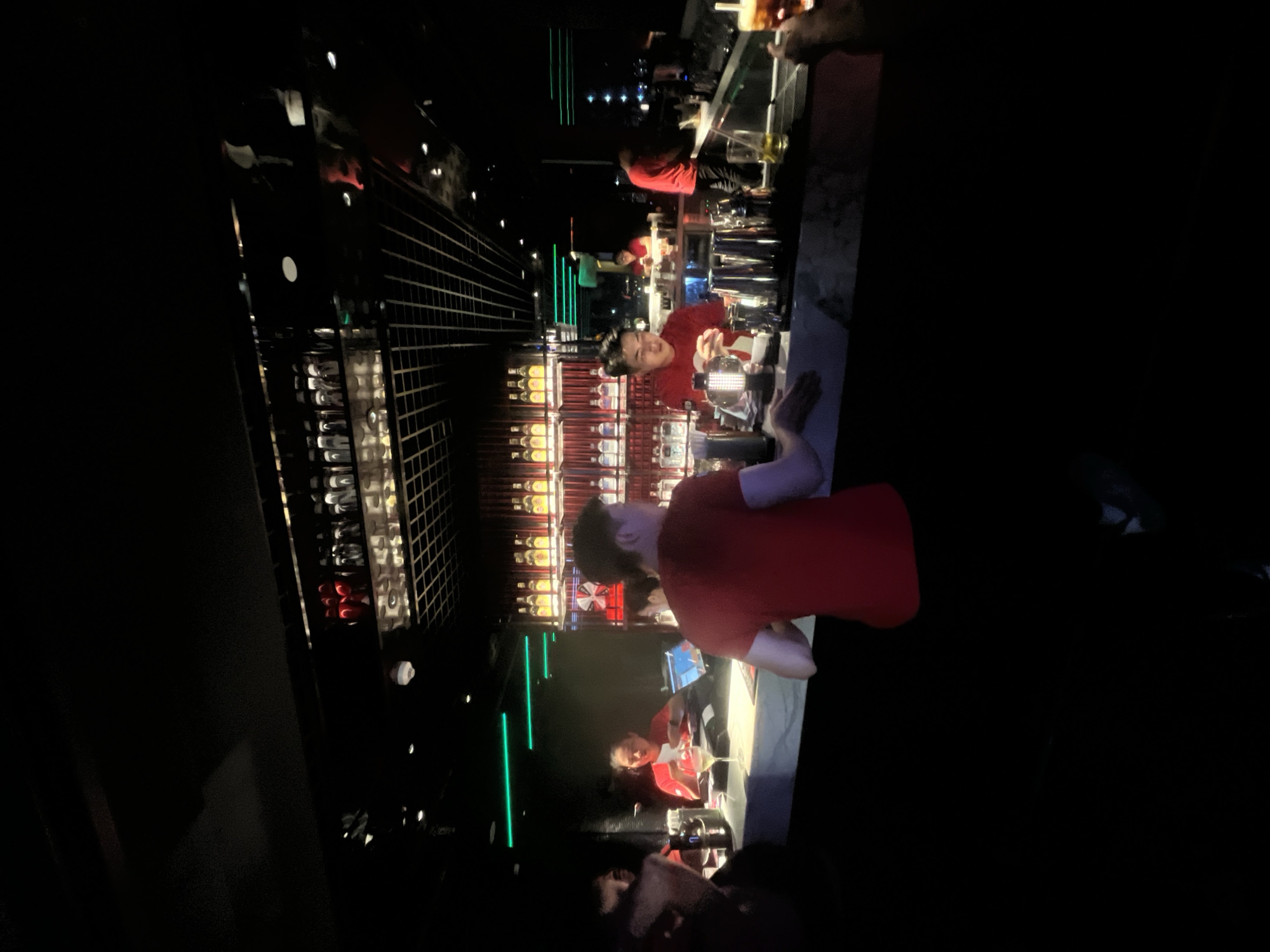 In the dimly lit interior of a bar in Đà Nẵng, Vietnam, patrons and staff interact around a polished bar. A bartender in a red shirt is visible on the left, facing a customer and holding a white card. Further down the bar, another bartender in a red shirt is actively preparing drinks, surrounded by various bar tools including cocktail shakers. Behind the bar, shelves are stocked with numerous bottles of alcohol, some illuminated by red and yellow lights, creating a vibrant display. The ceiling features a grid-like structure with downlights. Neon green lights are visible along the back wall. In the foreground, a patron on the far right holds a drink, suggesting a social gathering. The overall atmosphere is that of a lively nightlife establishment.