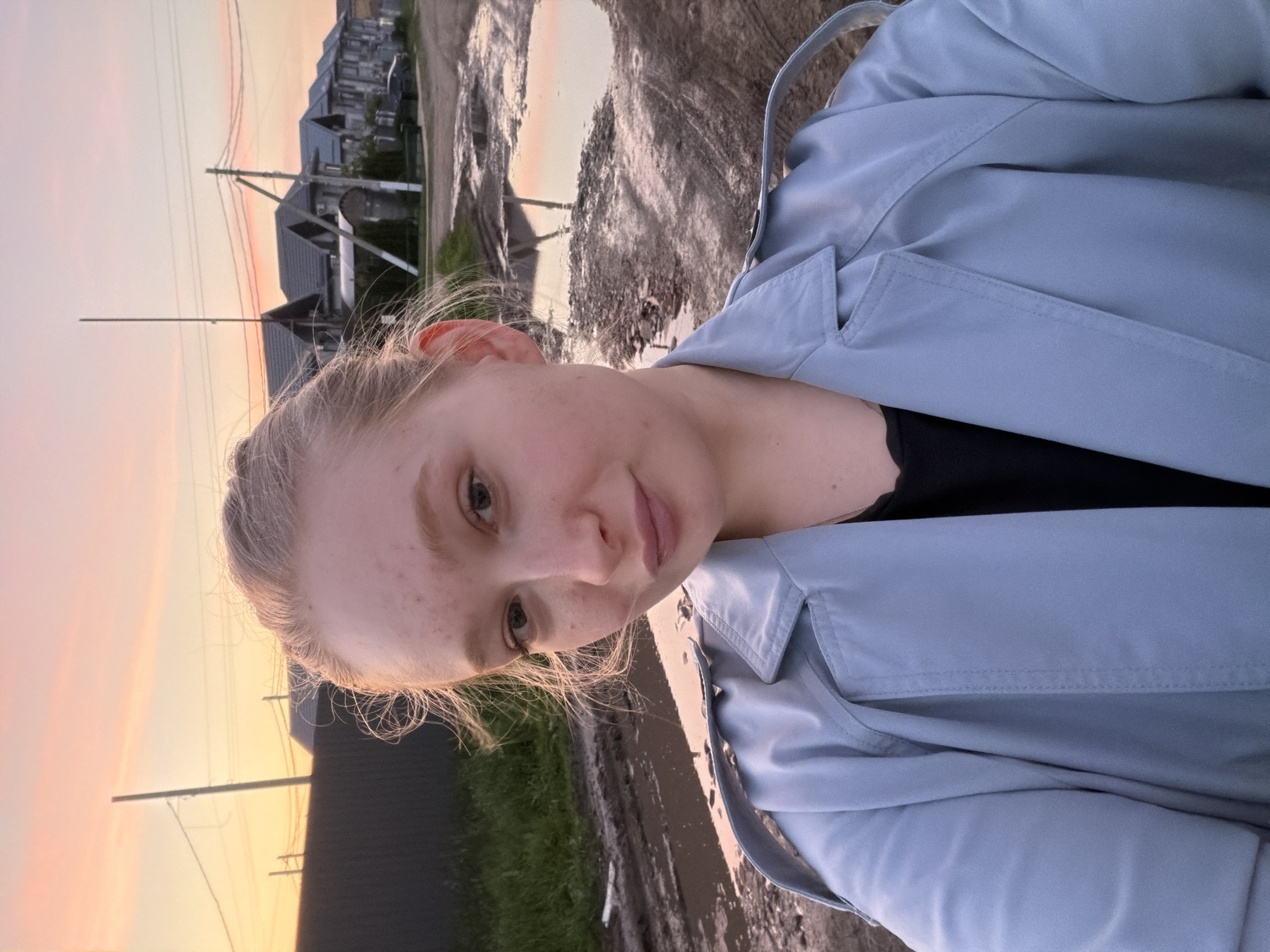A young woman is standing outdoors in Rossiyskiy, Russia. She is fair-skinned, with freckles, and her blonde hair is pulled back. She wears a light blue trench coat over a black top. The ground around her is muddy with puddles. In the background, there are houses, utility poles, and a sky with colors of orange and pink indicating either sunrise or sunset.