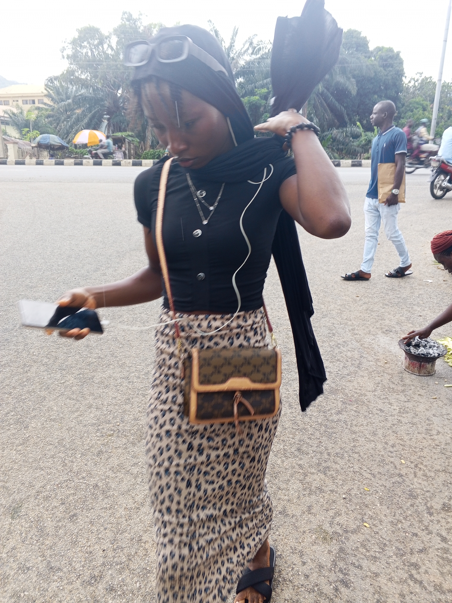 The image shows a woman in the foreground, wearing a black headscarf and a black t-shirt with a leopard print skirt. She is holding a smartphone in her left hand, which is plugged in, and a small brown handbag with a tan strap is slung across her body. She is wearing black sandals and a beaded bracelet on her right wrist. In the background, a man in a blue t-shirt and white pants is walking away from the camera, holding a brown folder. Another man is seated at a roadside stall with umbrellas. The scene appears to be a street in Lokoja, Nigeria, with palm trees and buildings visible in the distance. The ground is paved with asphalt.