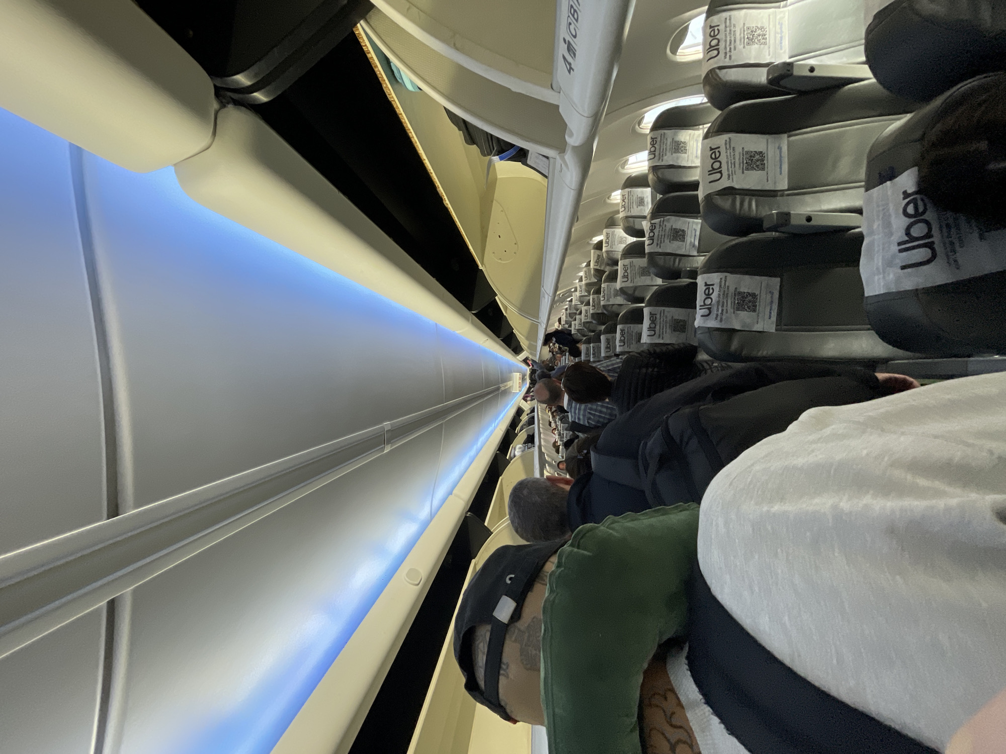 The image displays the interior of an airplane cabin, viewed from an aisle seat and rotated 90 degrees counter-clockwise. The perspective looks down a long aisle towards the front of the aircraft, with rows of passengers seated on both sides.
**Subjects:** Numerous passengers are visible, primarily from behind, occupying the dark grey or black seats. The foreground shows the back of a person wearing a green top and a black baseball cap, with tattoos visible on their arm. Other passengers' heads and upper bodies are discernible further down the aisle. The objects include the airplane seats, overhead bins, the cabin walls, and ceiling, illuminated by integrated mood lighting.
**Activity:** Passengers are settled in their seats, suggesting the plane is in the process of boarding, taxiing, or awaiting departure/arrival. There is no overt activity; the scene appears calm and orderly.
**Notable details:** A distinct blue mood light runs along the ceiling, casting a soft glow throughout the cabin. The seat headrests are covered with white protectors, each prominently featuring the "Uber" logo. Beneath the logo, QR codes are printed. One visible text accompanying a QR code explicitly states "valido por Argentina" (valid for Argentina) and hints at a promotion with Uber Eats. An overhead bin has the label "4M/CB". Given the location context of Ushuaia, Argentina, the "Uber" branding and the Spanish text align with this setting.
**Visible text:** "Uber" appears repeatedly on seat headrest covers. Text fragments "valido por Argentina" and "Uber Eats" are associated with QR codes. "4M/CB" is seen on an overhead bin.