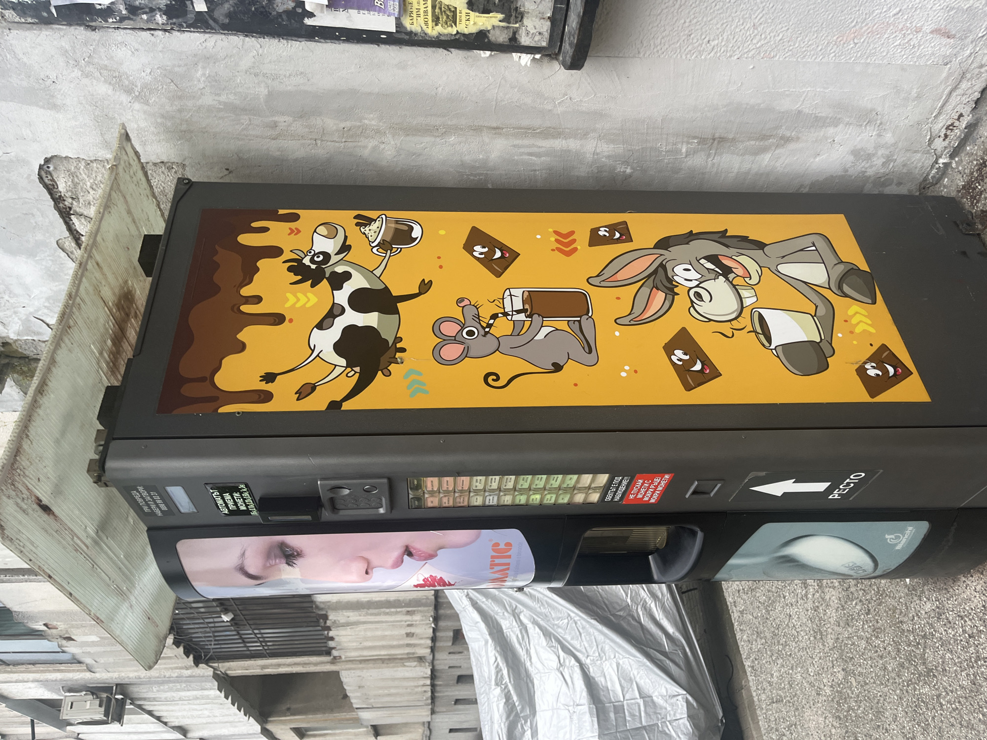 The image shows a vending machine, seemingly located outdoors. The top of the vending machine features a cartoon design with a cow, mouse, and donkey drinking beverages, and chocolate bars with faces, against a yellow background with brown liquid dripping down. The vending machine has selection buttons, slots for inserting money, and an area for product retrieval. There is also an advertisement of a female face on the machine. Writing on the machine appears to be in Bulgarian. The machine is labeled with "PESTO" and displays a service phone number. The environment around the machine includes parts of buildings, potentially rooftops or stacked items, and possibly a covering or tarp.