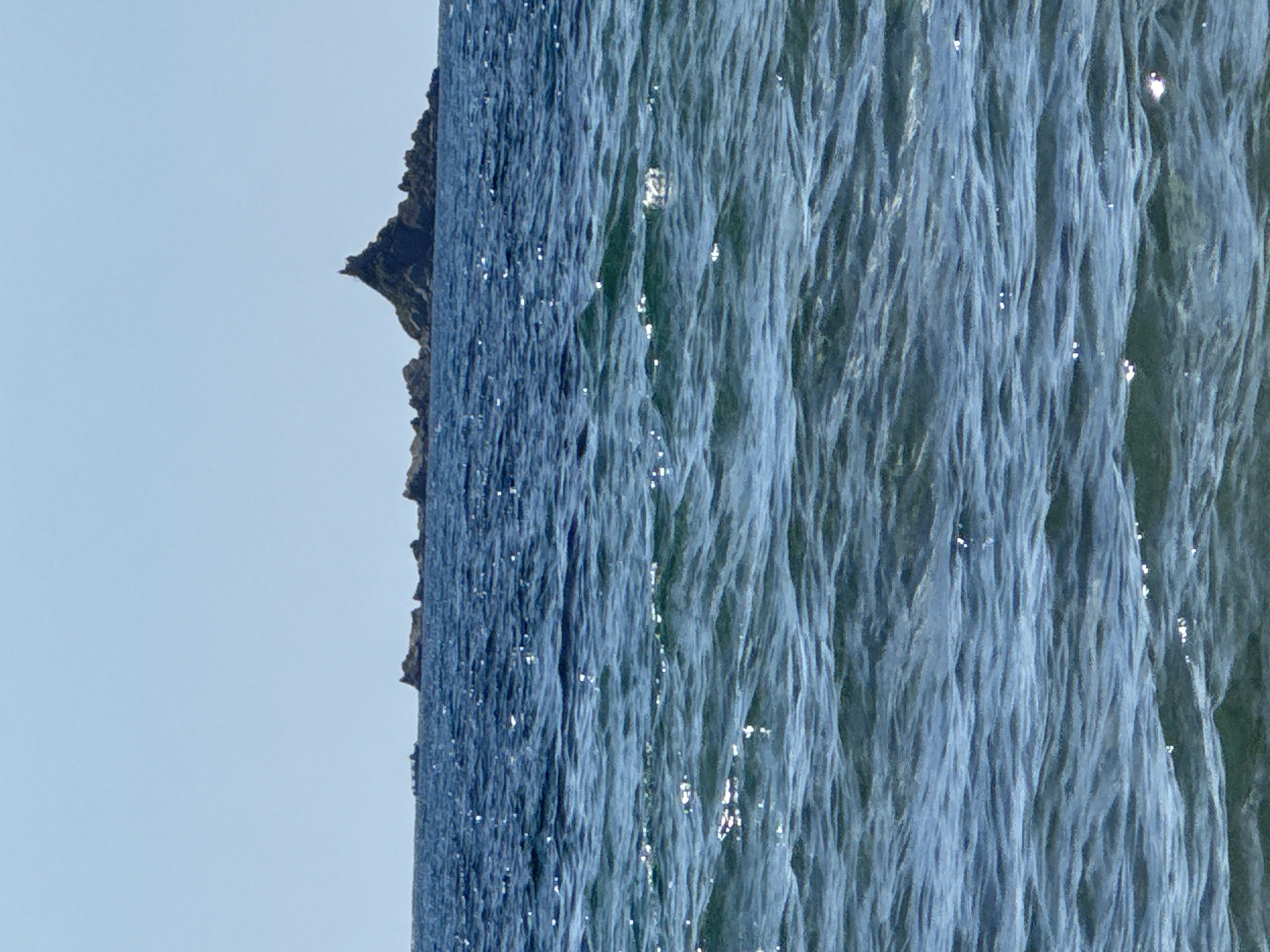 A distant view of a rocky island with a prominent peak and a structure atop it, set against a clear, pale blue sky. The island is surrounded by the deep blue sea, which exhibits moderate waves and reflections of sunlight. The image is captured from a distance, placing the viewer's perspective on the water. The scene is devoid of people or boats, presenting a natural, isolated seascape. The location is identified as being near Đurovići, Montenegro.