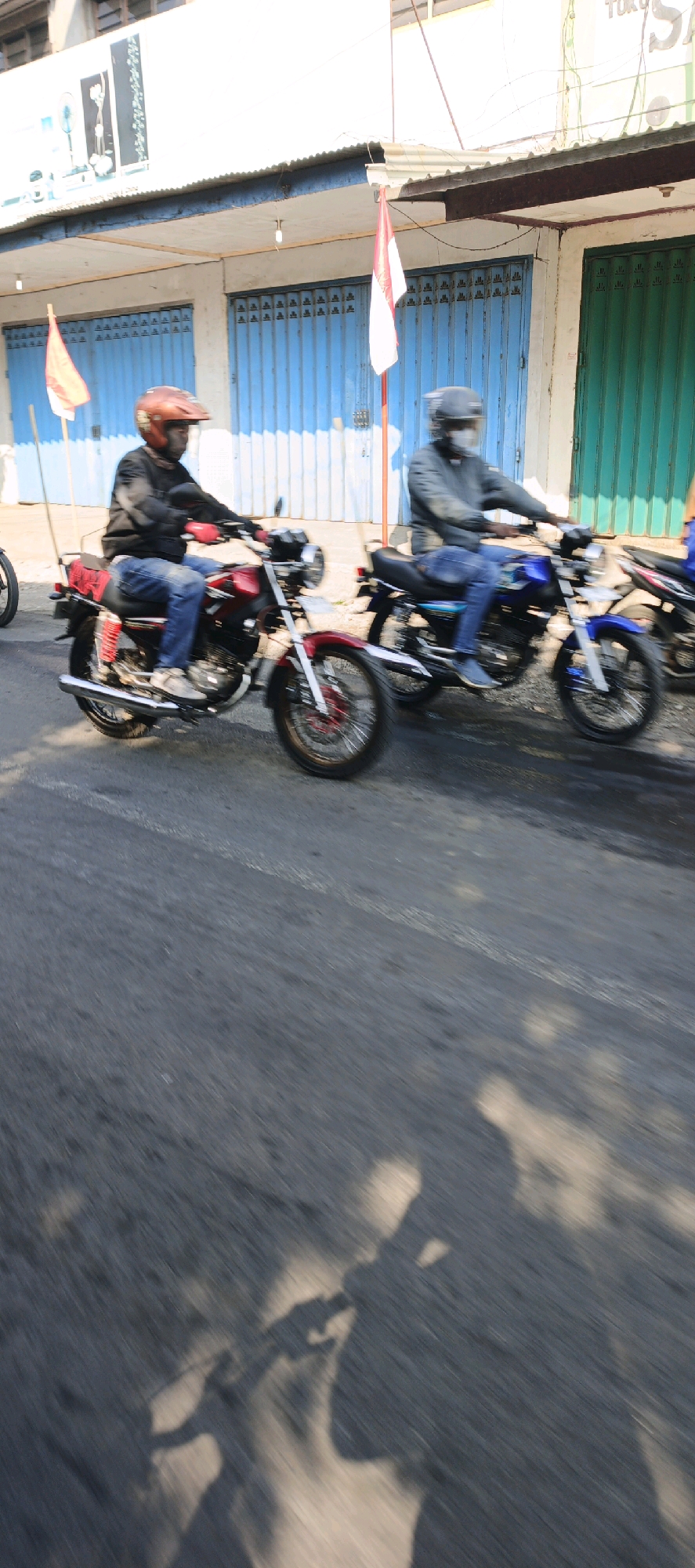 Two men on motorcycles are captured in motion on a paved road, likely in the city of Menampu, Indonesia. The motorcycle in the foreground is red and white, ridden by a person wearing a red helmet, black jacket, and blue jeans. The second motorcycle, partially visible to the right, is blue and is ridden by a person wearing a dark helmet and jacket, and blue jeans. They are riding past a building with blue and green metal roll-up doors. Indonesian flags are visible, suggesting a national holiday or celebration. The sunlight casts distinct shadows on the road. The image is blurred due to motion.