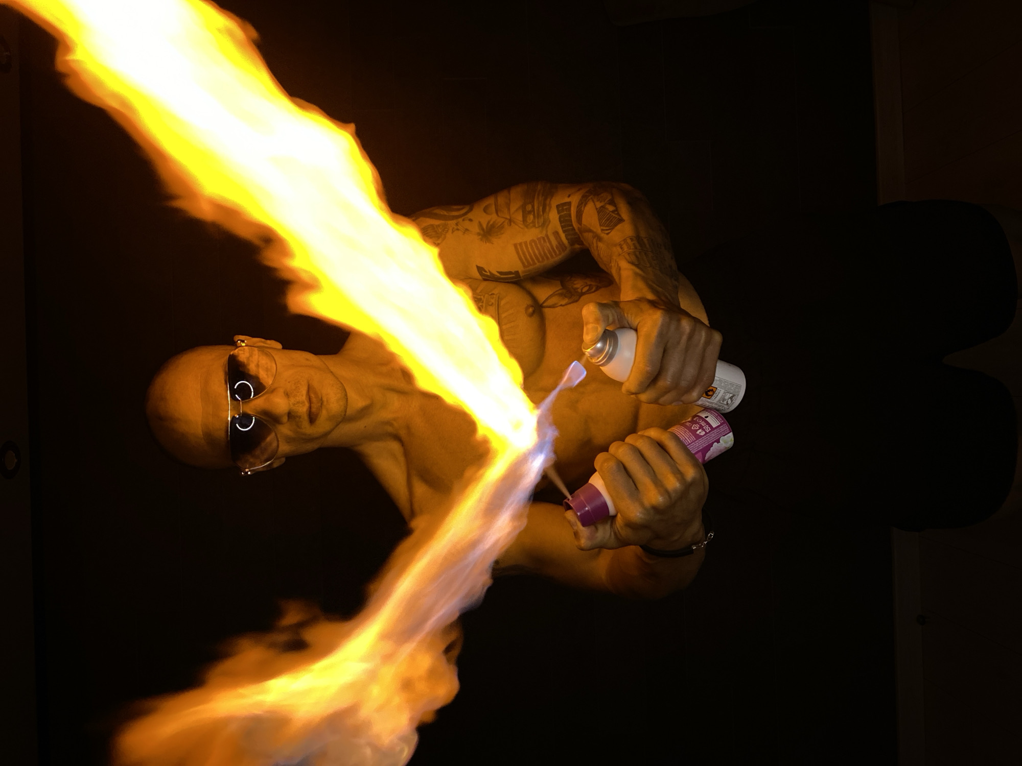 A shirtless, bald man wearing aviator sunglasses stands against a black background, holding two spray cans and creating a large flame. The man's left hand holds a purple and white spray can, while his right hand holds a white spray can. Both cans are spraying a substance that ignites into a bright yellow and orange flame, forming a V-shape in front of him. The man has tattoos on his left arm and chest. The scene appears to be indoors, with a dark, possibly unfinished wall or ceiling visible at the top. There is no discernible location in the city of Pleven, Bulgaria.