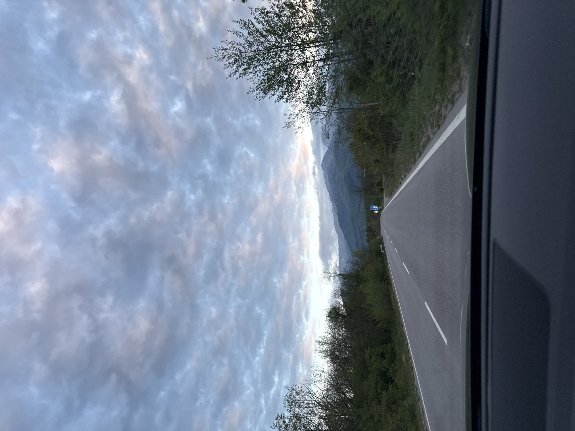 The image shows a view from inside a vehicle, likely a car, looking out onto a road in a mountainous area. The road has a single white line marking the lane. Trees line both sides of the road, and in the distance, there are visible mountains partially obscured by clouds. The sky is filled with clouds, and the lighting suggests it is either dawn or dusk. The location is possibly near Mezdreya, Bulgaria, based on the environmental characteristics.