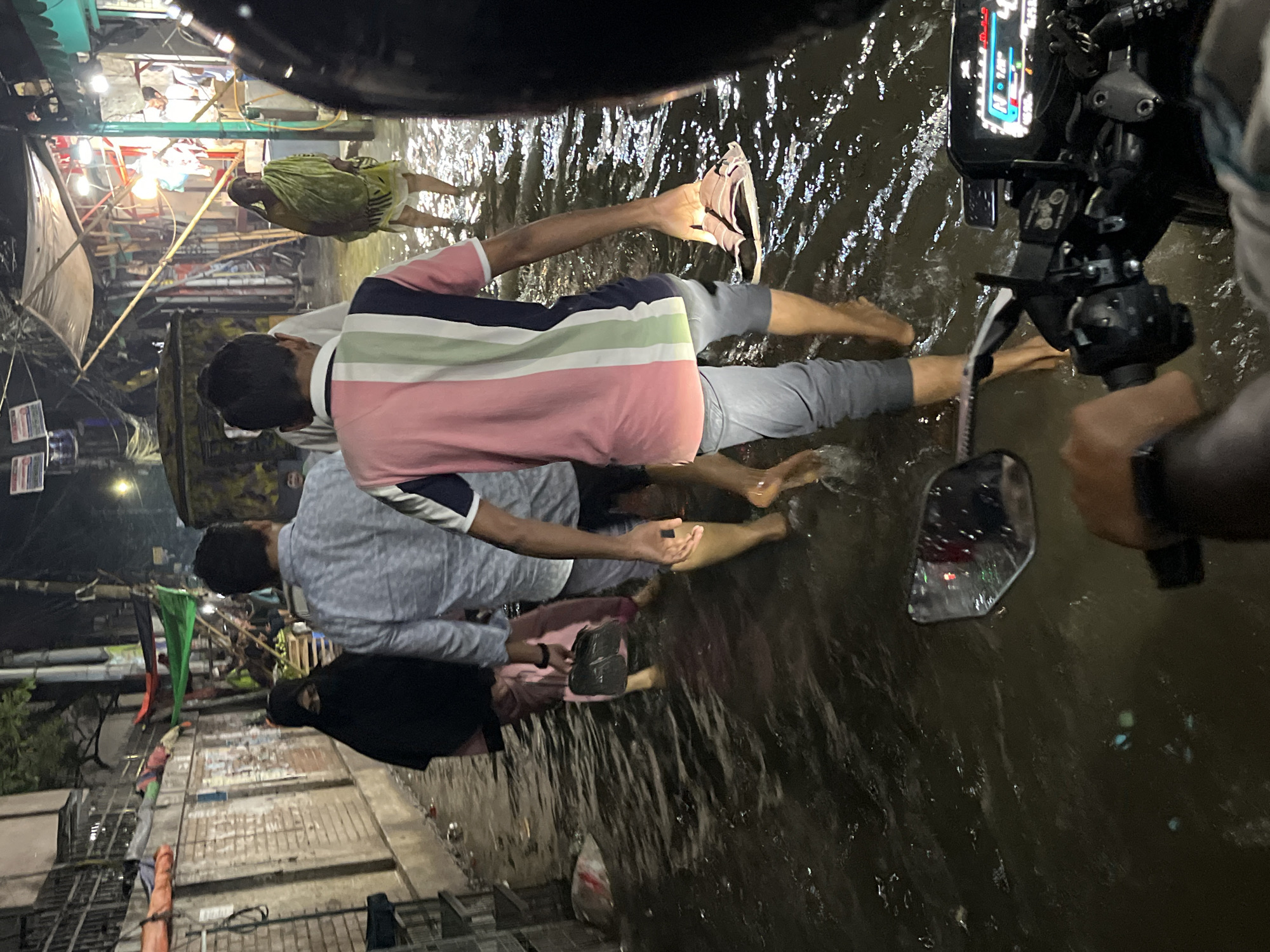 The image depicts a flooded street scene in Dhaka, Bangladesh. The floodwater reaches the knees of pedestrians wading through it. Several individuals are visible, including a person in a pink and white striped shirt holding a sandal in the water, a person wearing a gray shirt behind them, and a woman in a black burqa. The water reflects the lights of shops and buildings on either side of the street. A motorcycle is in the right foreground, partially submerged in the water. The overall environment suggests a crowded urban area experiencing significant flooding.