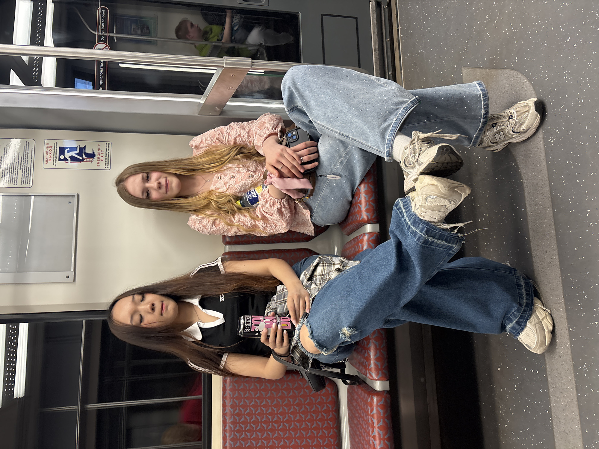 The image depicts the interior of a subway train car, identified as part of the Saint Petersburg Metro in Russia by the Cyrillic and English signage ("АВАРИЙНЫЙ ВЫХОД / EMERGENCY EXIT," "НЕ ПРИСЛОНЯТЬСЯ / Do not lean on door!"). Two young women are seated on red-patterned seats, facing forward.
The woman on the left has long, dark brown hair, closed eyes, and an expression suggesting introspection or rest. She wears a black polo shirt with white trim on the collar and sleeves, ripped blue jeans, and light-colored chunky sneakers. She holds a pink and black "BERRYCRU MAX ENERGY PINK" beverage can in her hands, which are resting on a folded plaid shirt draped across her lap. A black shoulder bag is visible by her side.
The woman on the right has long, blonde hair and is looking directly at the camera with a slight smile. She wears a pink, textured long-sleeved blouse, light blue jeans adorned with small sparkly embellishments, and light-colored chunky sneakers (one with visible "N" branding, likely New Balance). She holds a dark-colored smartphone and a pink scrunchie. A small "Aqua" brand water bottle is visible next to her.
Both women have their legs crossed. In the reflection of the train door to the right, a young male figure in a green shirt is partially visible, seated across from the women. The train car features white walls, dark windows, metallic fixtures, and a grey floor with white speckles.