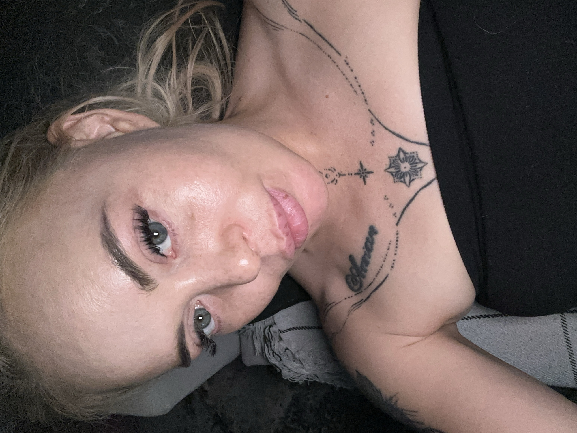 An adult female is depicted from an overhead perspective, looking directly into the camera. She has fair skin, blonde hair, blue-gray eyes with prominent eyelashes, and defined eyebrows. Her lips are full and slightly parted in a pout. Her skin exhibits visible pores, a slight sheen, and some minor textural variations on the forehead, nose, and cheeks, with a few small freckles present. She is wearing a black strapless garment. Multiple tattoos are visible on her upper chest and right collarbone: a script reading "Shania" (or similar) on the right collarbone, and intricate linear patterns featuring stars and dots centered on her sternum, extending towards both shoulders. A darker, indistinct tattoo is partially visible on her left upper arm. The background on the left consists of a textured gray fabric, possibly a blanket or cushion, with a striped pattern. The area behind her head and to the right is dark and largely undifferentiated, suggesting an interior setting with subdued lighting. The image depicts a scene within an interior space in the city of Unknown, Unknown.