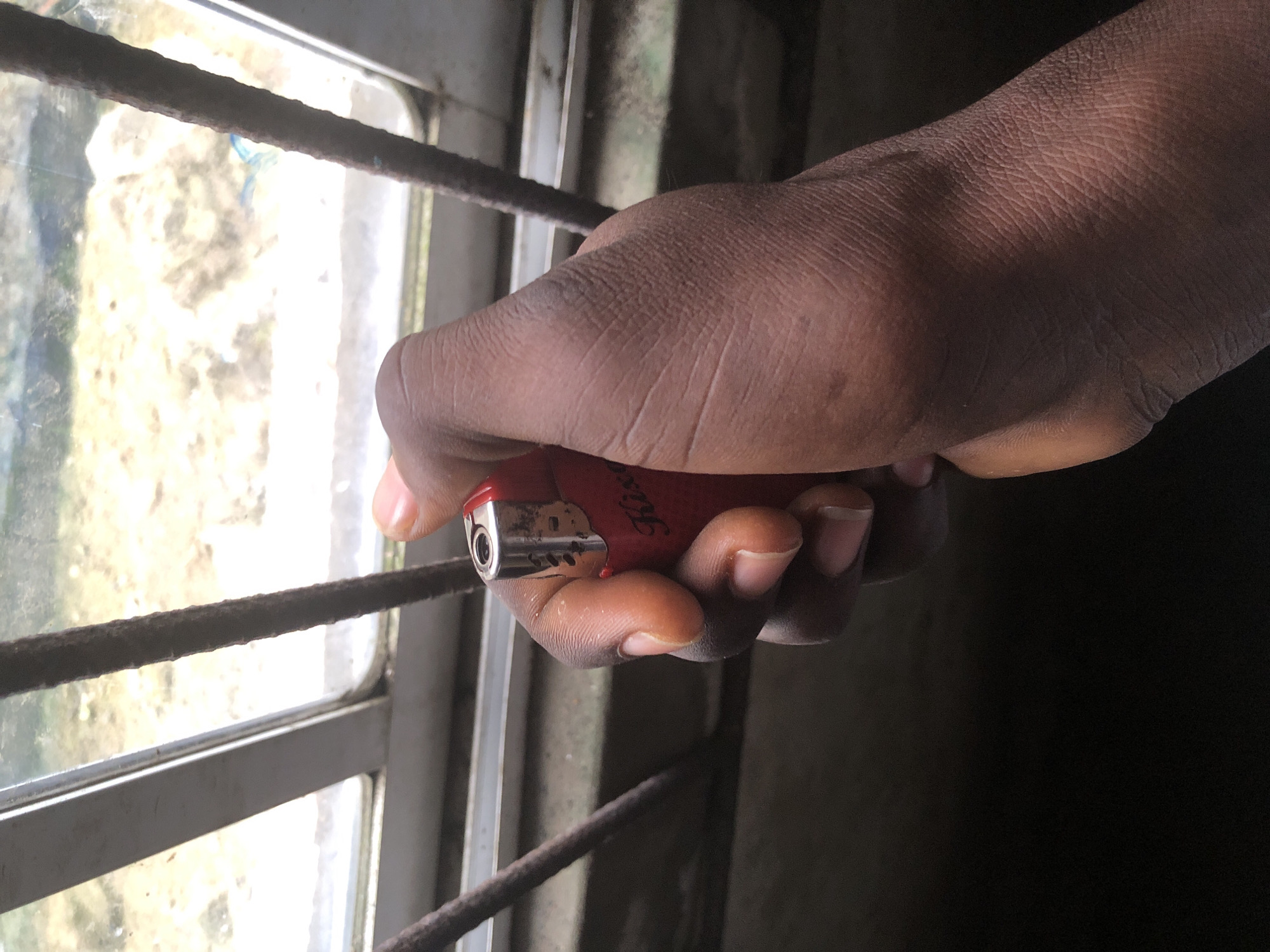 A close-up, upward-angled shot shows a dark-skinned person's hand holding a red disposable lighter. The lighter has a silver tip and a label with red lettering that reads "His...". The hand is positioned in front of a window with horizontal metal bars. The window pane is dirty and looks out onto a blurry, sunlit, outdoor scene that appears to be an unpaved area with some sparse vegetation. The scene is dark with heavy shadows, suggesting it is either indoors or taken during a time of low light. The presence of the metal bars and the window suggests an urban or semi-urban setting. Given the prompt's request for a location in Ado, Nigeria, this could be a view from a room or structure with bars on the window, possibly for security or ventilation.