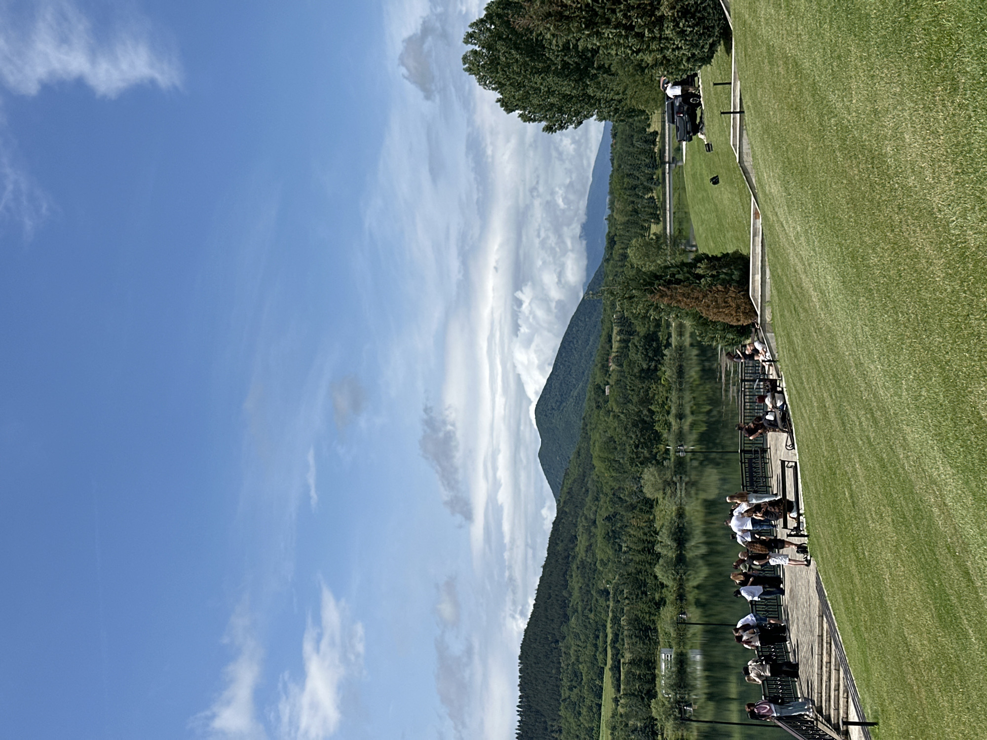 The image captures a serene outdoor scene at Razliv, Bulgaria, depicting a wide expanse of water bordered by lush green landscapes and mountains under a bright sky. The photograph is oriented vertically, with the horizon running down the center, presenting a panoramic view.
In the foreground, on the right side of the frame, there is a meticulously maintained green lawn or golf course. Adjacent to this grassy area is a paved walkway or patio, protected by a dark railing. Along this railing, several individuals are gathered. Most are adults, casually dressed, either standing and observing the scenic view or seated on benches, seemingly socializing or relaxing. A golf cart or similar utility vehicle is visible on the grass further up.
The left side of the image features a calm body of water, likely the Razliv Reservoir, which clearly reflects the sky and the dense forest along its banks. Beyond the water, a range of forested mountains rises, their peaks appearing in the distance under a slightly hazy sky. The mountains are covered in thick greenery, indicating a rich, natural environment.
The sky above is a clear, vibrant blue, adorned with scattered white clouds, some appearing wispy and elongated. The bright daylight suggests it is late morning or early afternoon, with excellent visibility and pleasant weather conditions. The scene evokes a sense of tranquility and leisure, with people enjoying the picturesque natural surroundings. There is no visible text in the image.