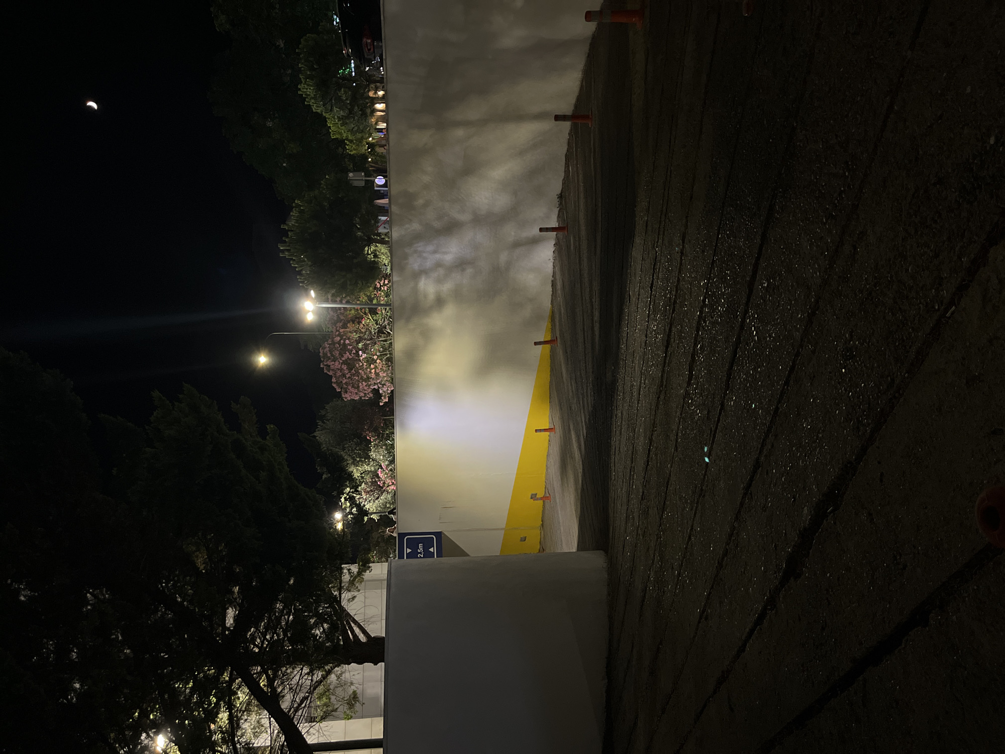 The image shows an outdoor view at night, likely in Budva, Montenegro, focusing on a ramp or entrance to an underground area. The ground in the foreground is dark and appears to be asphalt or a similar rough surface, with visible tire tracks and possibly some moisture. Several orange traffic cones and bollards are placed along the edge of the ramp, guiding the path.
To the left, a curved, white concrete wall forms the entrance, with a blue sign indicating "2,5m" which likely refers to a height restriction. Beyond this wall, more of the ramp is visible, illuminated by artificial lighting. This lighting casts shadows on the white wall, creating a textured effect.
In the background, a row of trees and flowering bushes line the top of a taller, illuminated wall. Streetlights are visible among the trees. The sky is dark, with a sliver of a crescent moon visible in the upper right corner. The overall impression is of a service entrance or parking area at night. There are no people clearly visible in the immediate scene.