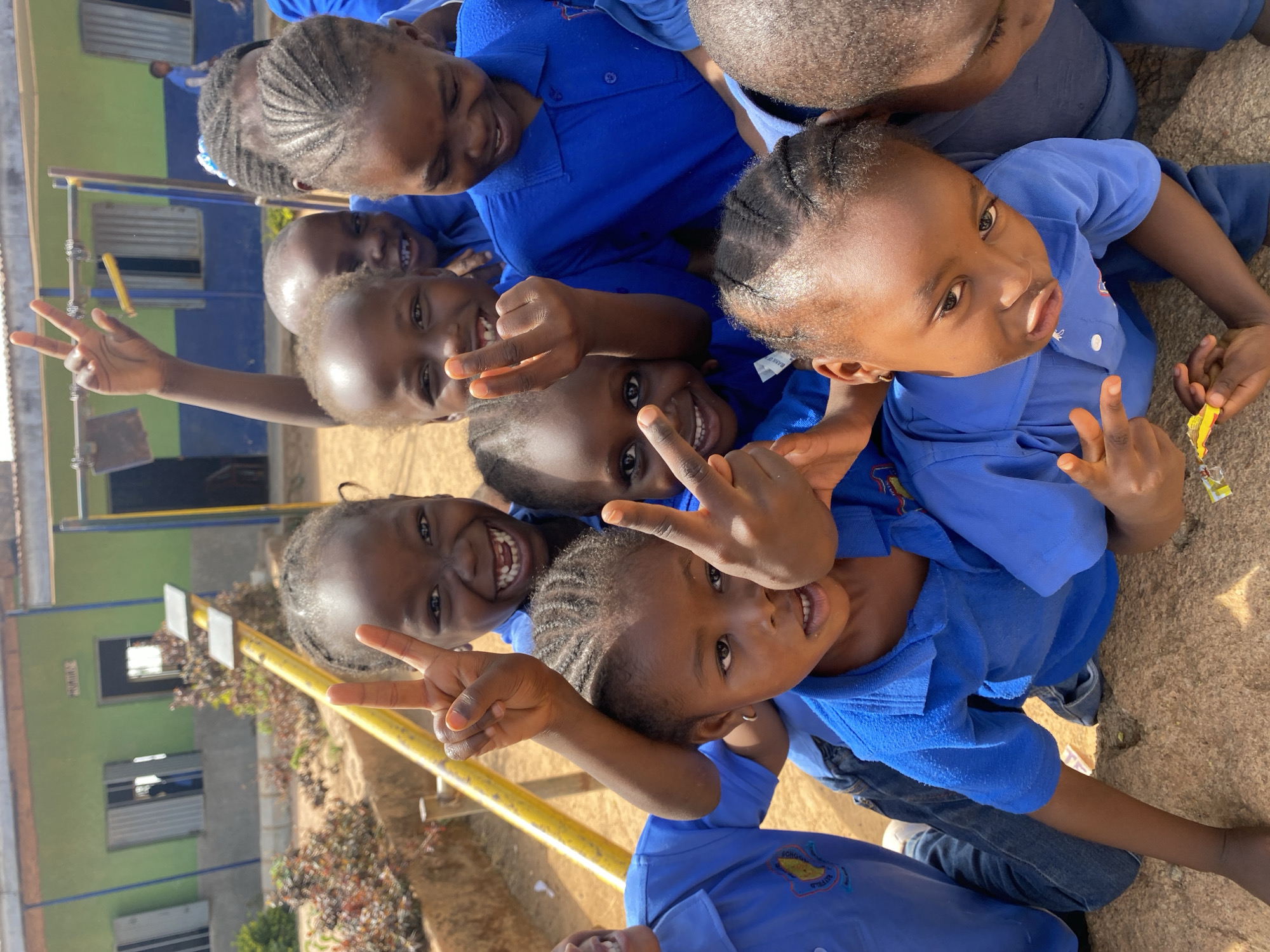 A group of at least nine Black children, all wearing blue collared shirts, are crowded together, looking up at the camera, several with their hands raised making a "peace" or "victory" sign. The children appear to be of primary school age. In the background, a building with a yellow bar is visible. The location is Gura Top, Nigeria, based on the shirt label. The ground appears to be sandy or dirt-covered.