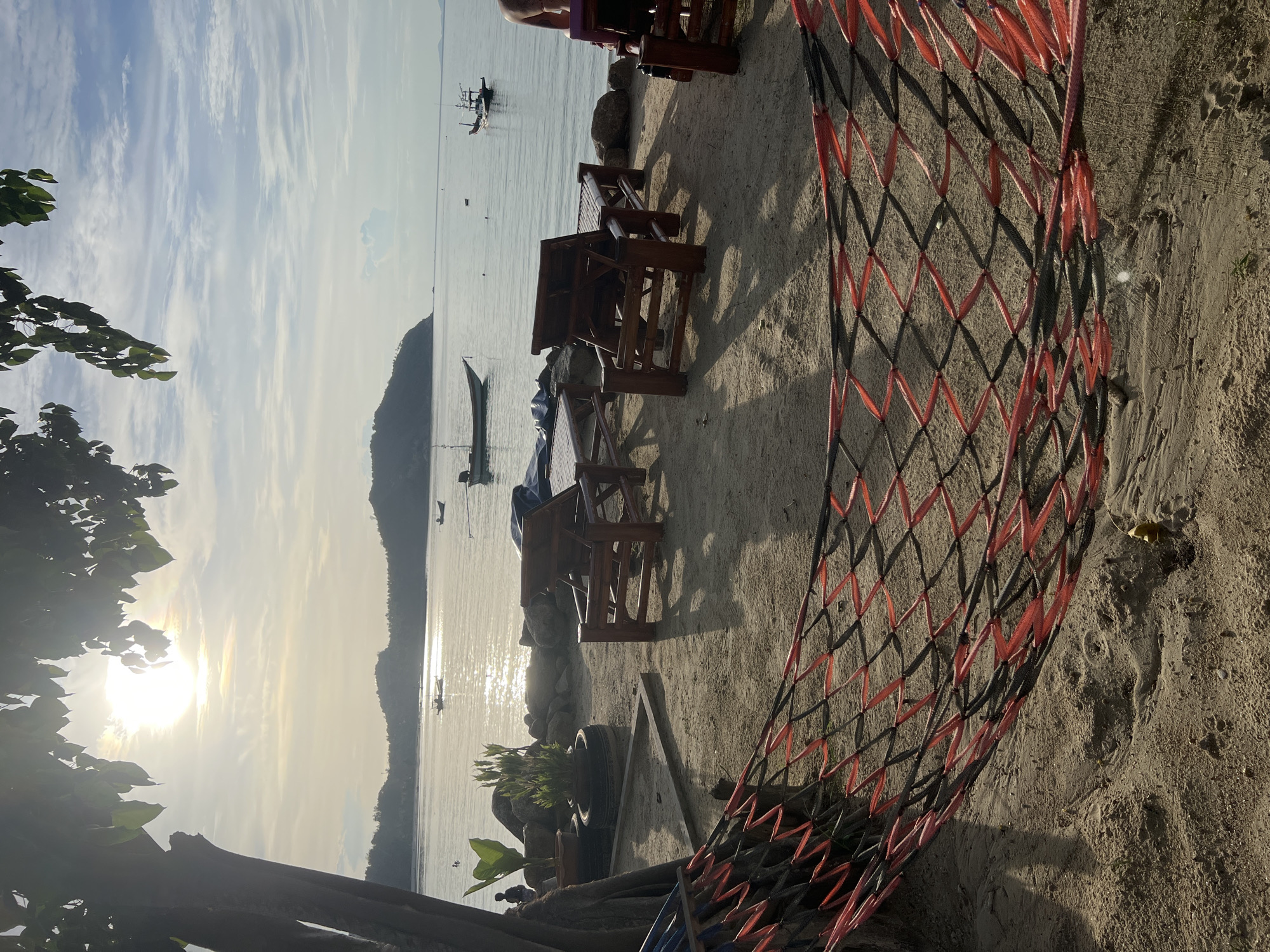 The image shows a serene beach scene at sunset. In the foreground, a red and black hammock is strung across the sand, with its diamond pattern casting shadows. Behind the hammock, several wooden tables and chairs are set up on the beach, suggesting an outdoor seating area for a restaurant or cafe. The sand is light brown and appears to be slightly damp in places.
The middle ground features the calm ocean water, with several small boats anchored offshore. The sun, low in the sky, casts a warm, golden glow across the water, creating a shimmering effect. A hazy, mountainous island is visible in the distance, silhouetted against the bright sky.
The sky is a mix of blue and white, with streaks of clouds that catch the sunlight. Lush green foliage from trees frames the left side of the image, adding a touch of nature to the scene. The overall mood is peaceful and relaxing, evoking a sense of tranquility and a tropical vacation. The lighting suggests it is late afternoon or early evening, with the sun beginning to set.