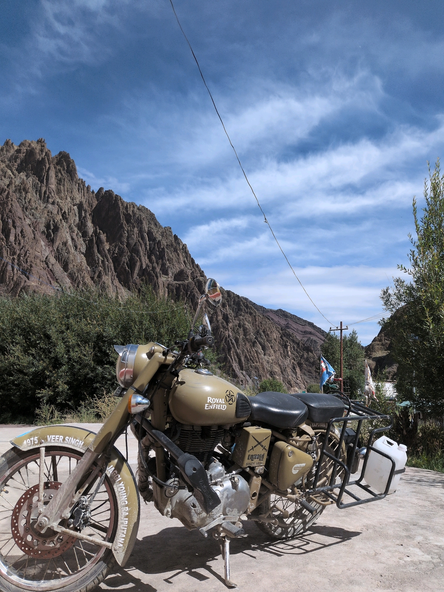 A stationary, tan-colored Royal Enfield motorcycle is positioned on a paved surface in front of a rocky, mountainous landscape under a partly cloudy blue sky. The motorcycle, facing slightly left, has a spare fuel can attached to its rear rack. Text is visible on the front fender, reading "1975 VEER SINGH," and on the side of the motorcycle. Flags are visible in the background, one of which is a blue and white flag. Green foliage is present both in front of and behind the motorcycle, with trees on the right side of the frame. The scene appears to be located in Lato, India, given the presence of the motorcycle with an Indian registration and the general landscape characteristics often associated with the region.