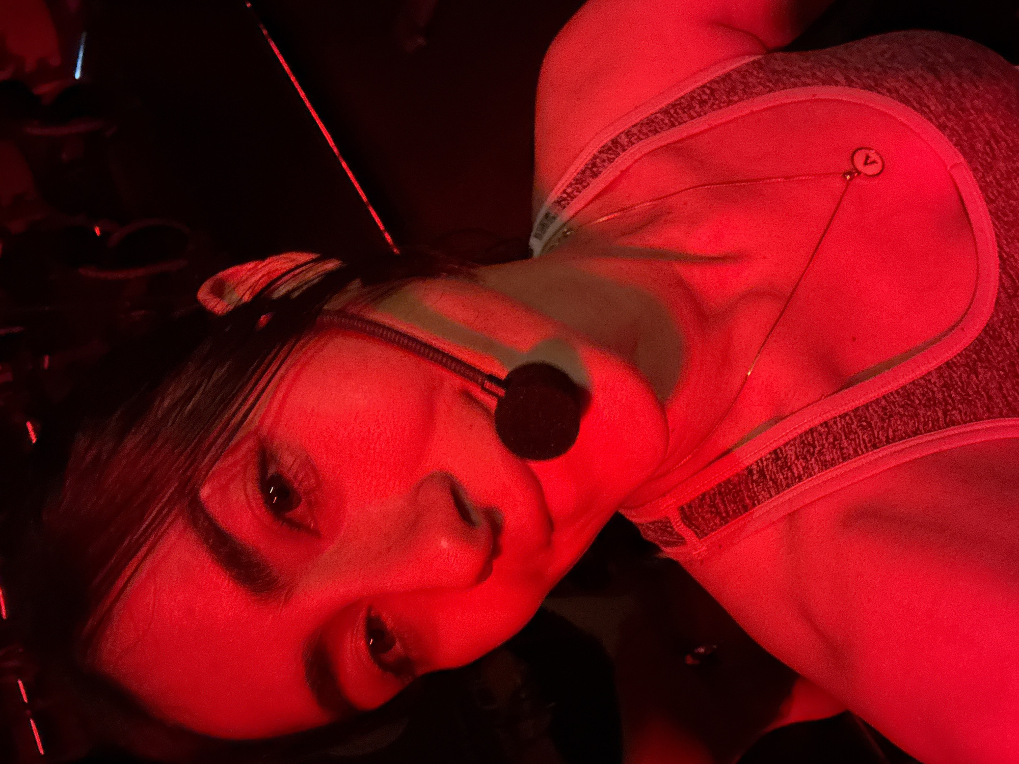 A woman is positioned in a prone position, illuminated by intense red lighting. She is wearing a grey tank top with a decorated edge and a microphone headset. An adhesive patch with a "7" printed on it is affixed to her upper chest. The background is dark and unclear. There is no discernible evidence indicating the location as Puebla, Mexico.