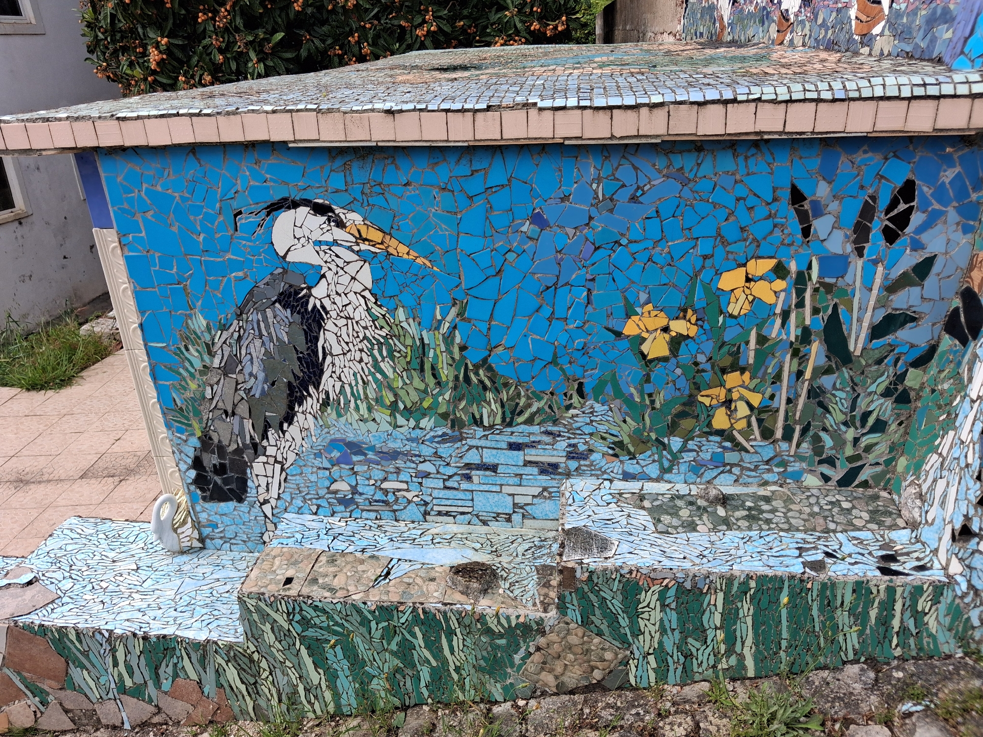 A mosaic-covered structure stands outdoors in Soure, Portugal. The dominant feature of the artwork is a depiction of a gray heron, rendered in fragmented tiles, set against a blue background. Additional details include stylized yellow flowers and green vegetation, also composed of mosaic pieces. The structure has multiple tiers, each adorned with mosaic designs that visually represent water and foliage. The overall impression is a decorative, outdoor installation, likely a public art piece.