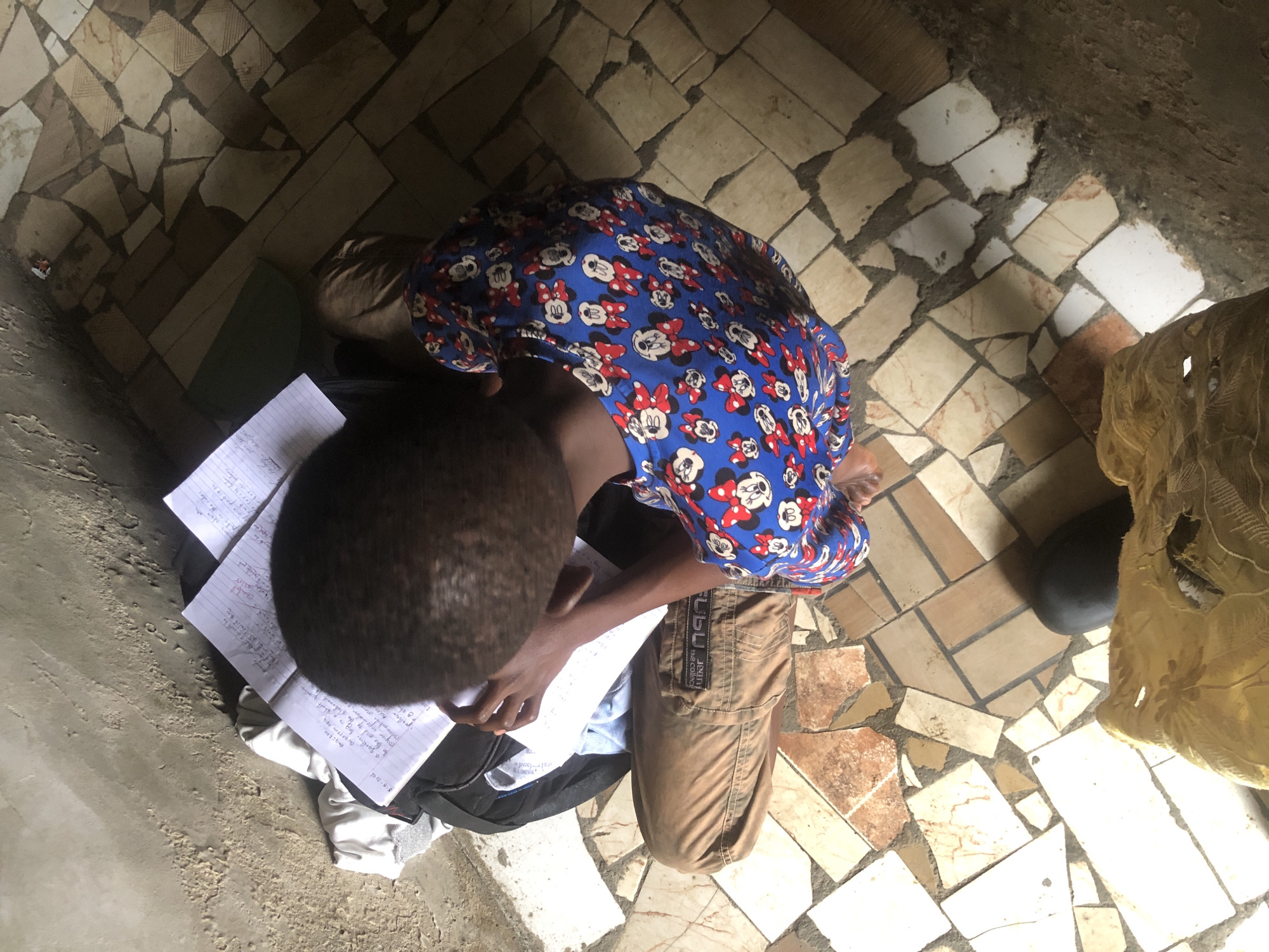 In Ado, Nigeria, a young boy is seated on the floor, engrossed in his studies. He is wearing a blue t-shirt with a Mickey Mouse pattern and brown cargo pants. The boy is surrounded by notebooks and papers, suggesting a dedicated study session. The floor is tiled with a mosaic of brown, white, and pinkish tiles, and a portion of a yellow blanket is visible in the foreground. The overall scene depicts a studious moment in a simple, tiled room.