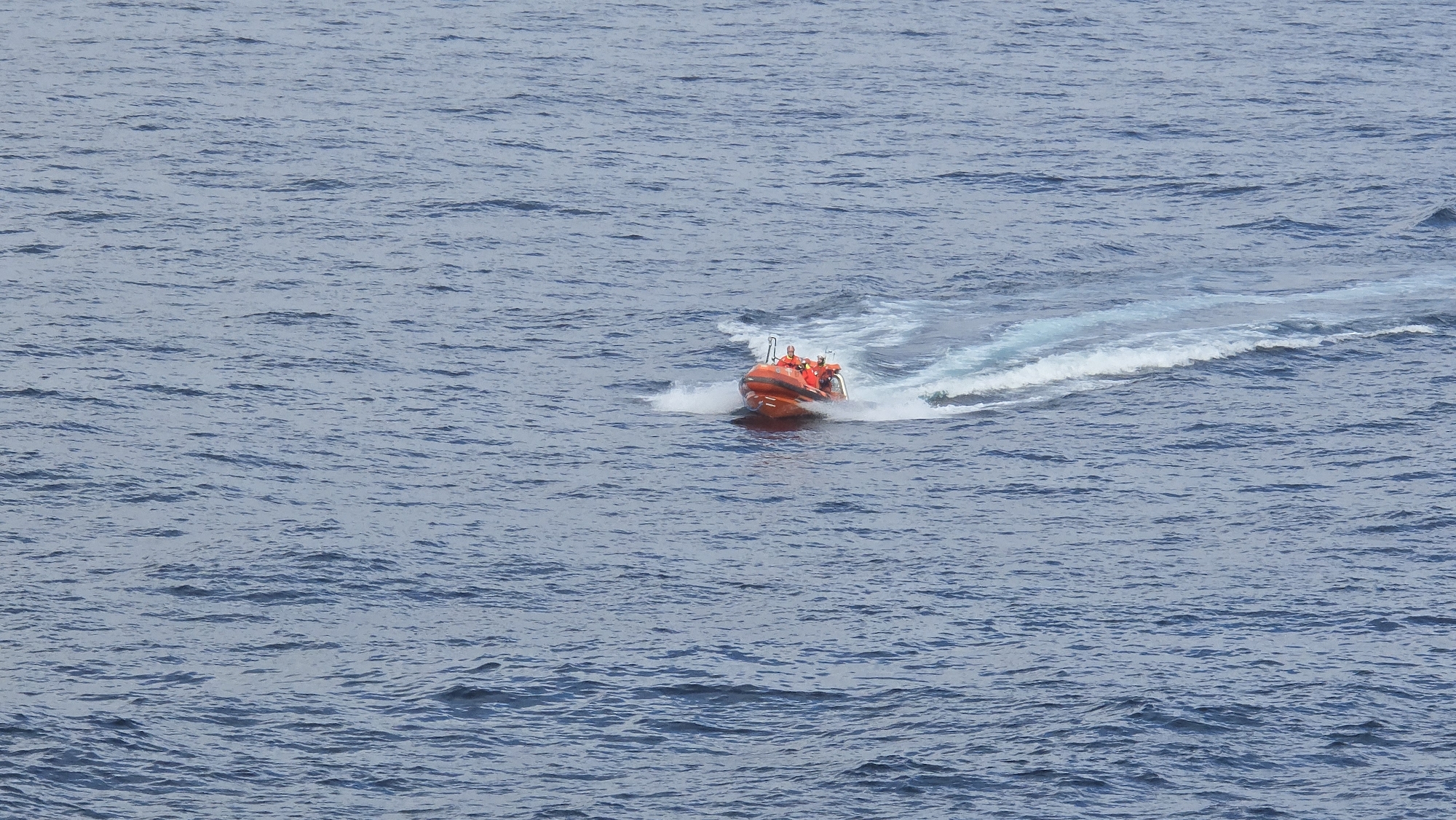The image depicts an orange inflatable boat moving rapidly across water. The boat has multiple occupants dressed in similar orange attire. The boat is creating a white wake in the water behind it. The location is on a body of water, and the city and region are Unknown, Unknown.